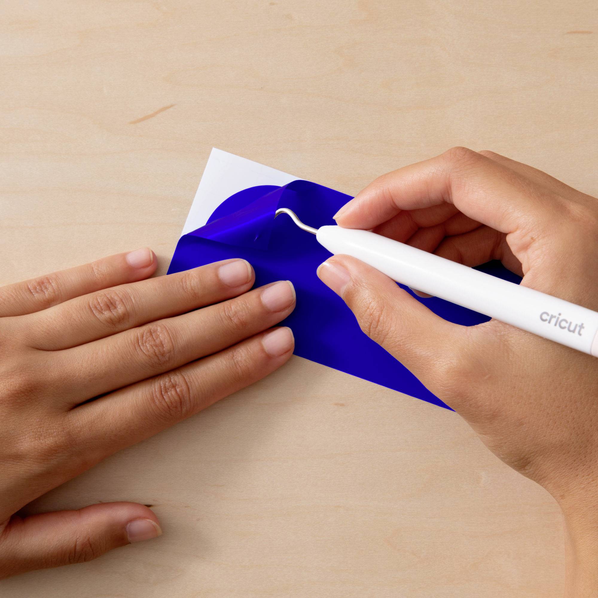 Ein Cricut-Werkzeug wird verwendet, um ein blaues Vinylmotiv auf einer Holzoberfläche zu entgittern. Hände führen den Arbeitsvorgang aus.