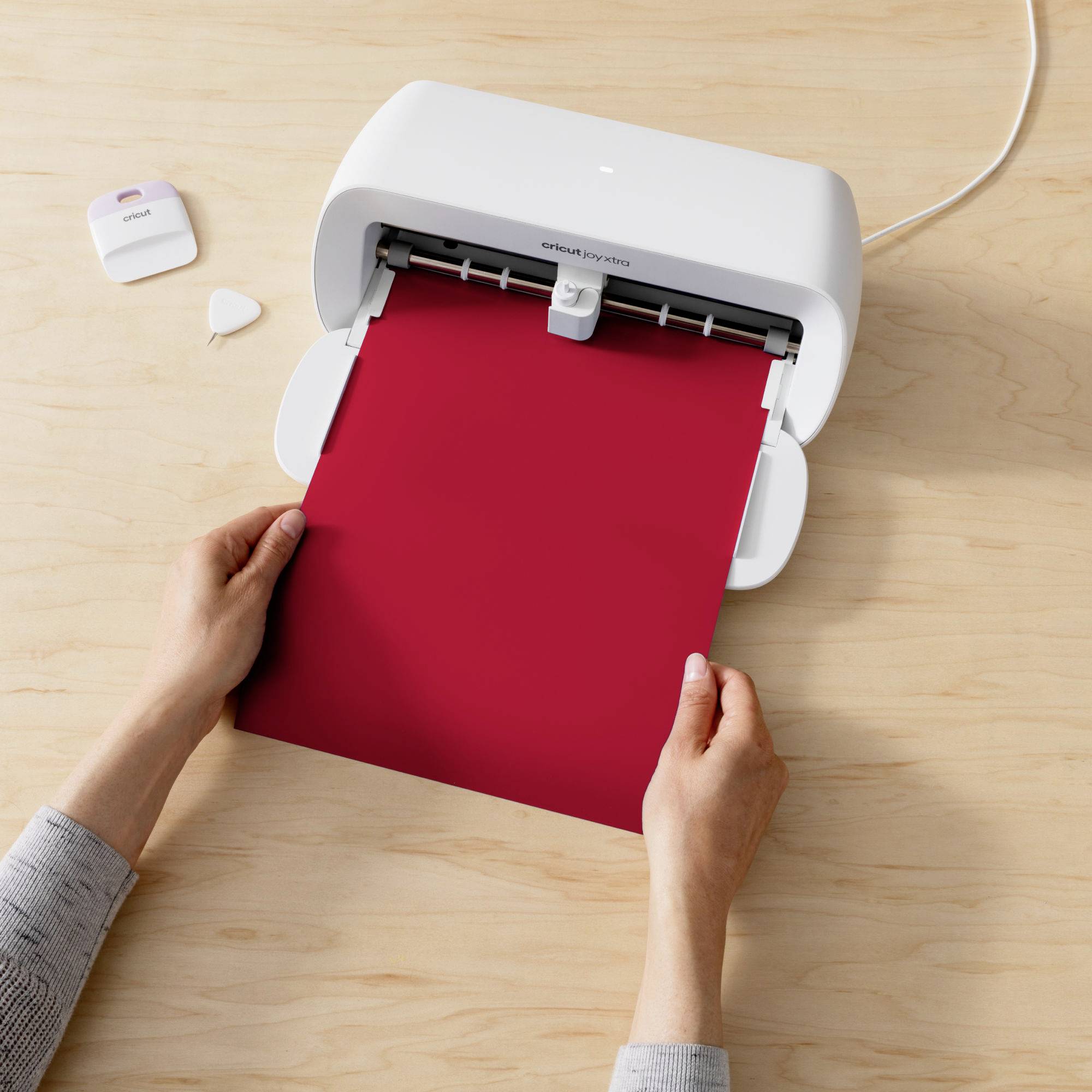 Person hält rotes Papier in einem Schneideplotter auf einem Holztisch, bereit zum Schneiden. Gerät im Vordergrund, Zubehör links.