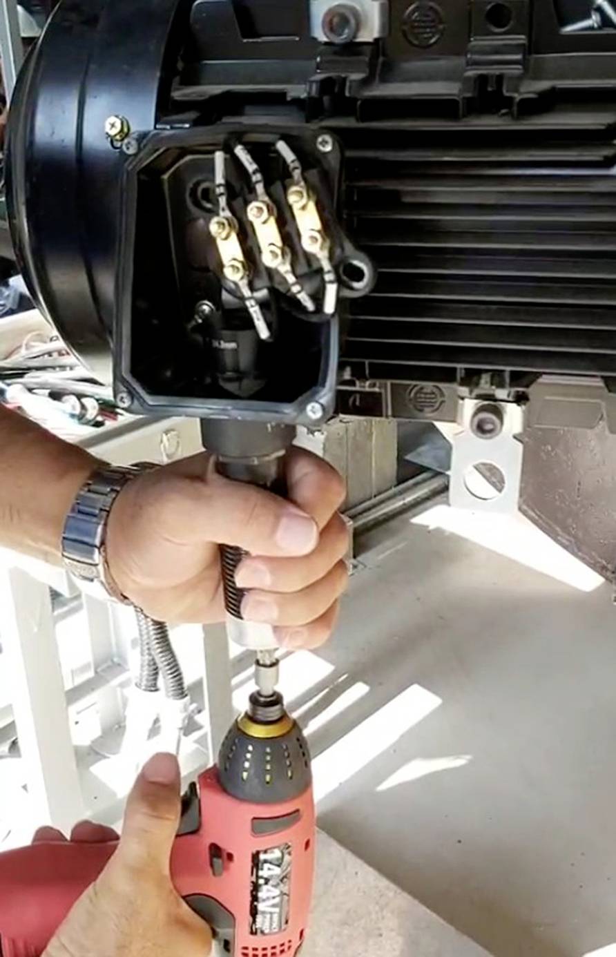 Eine Person arbeitet mit einem roten Elektrobohrer an der Wartung eines Elektromotors. Es sind verschiedene Kabel und Schrauben sichtbar.