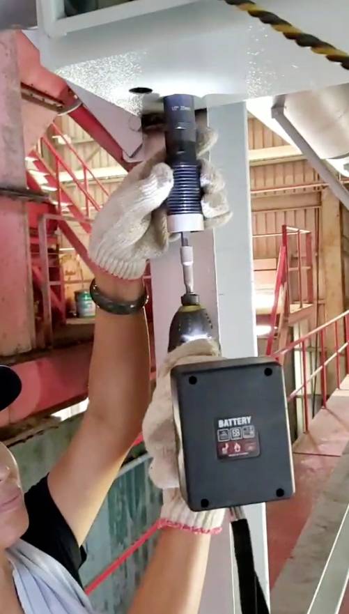 Eine Person in Handschuhen benutzt einen Akku-Bohrschrauber, um eine Schraube in ein Metallgerüst in einer industriellen Umgebung zu drehen.