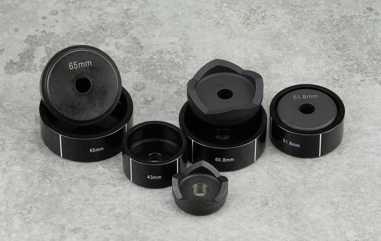 Eine Sammlung von schwarzen Metallhülsen in unterschiedlichen Größen liegt auf grauem Beton. Größenangaben (51,8 mm, 43 mm, 65 mm) sind sichtbar.