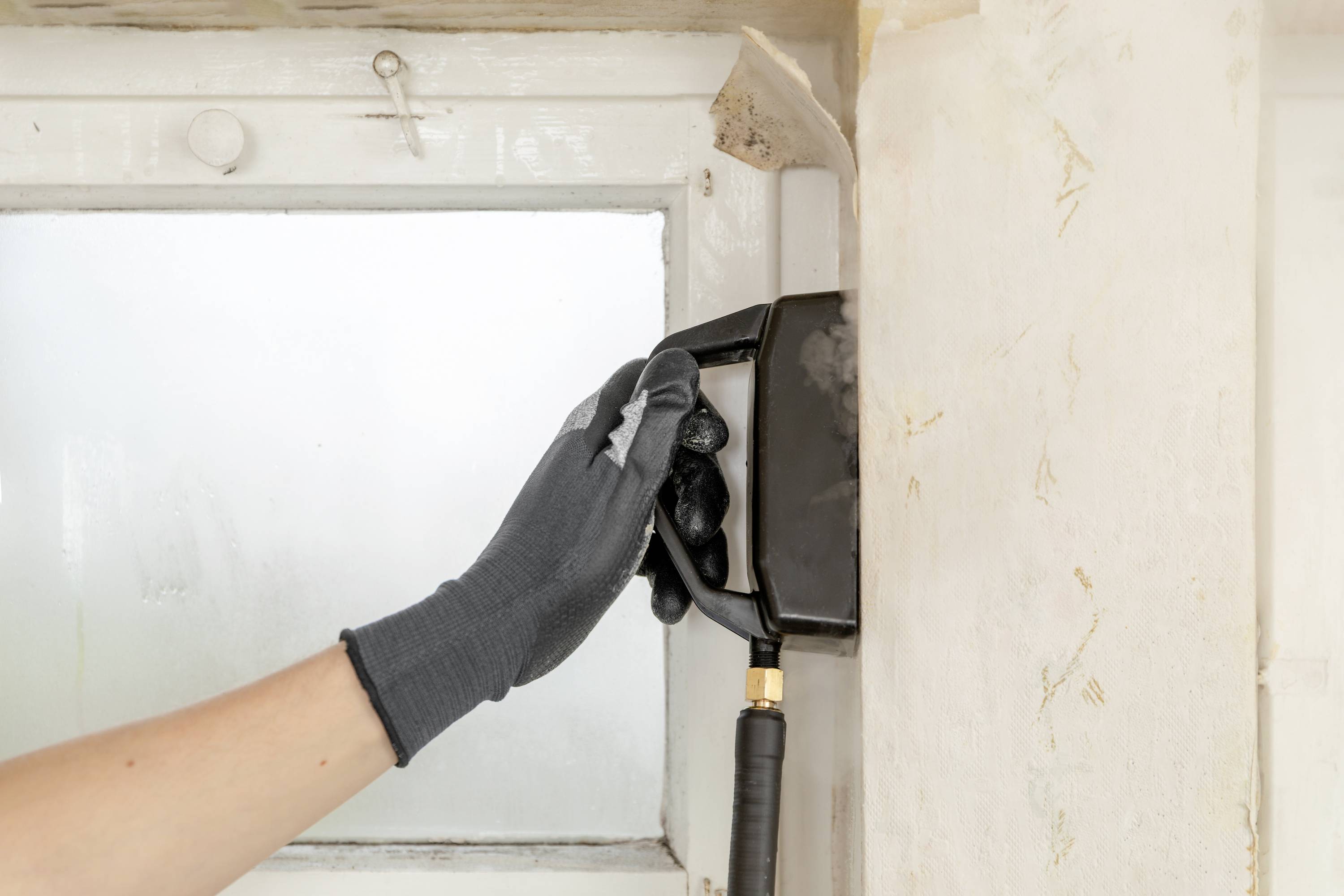 Eine Hand mit Handschuh betätigt einen Fensteröffner. Fensterrahmen und Wand sind leicht abgenutzt.