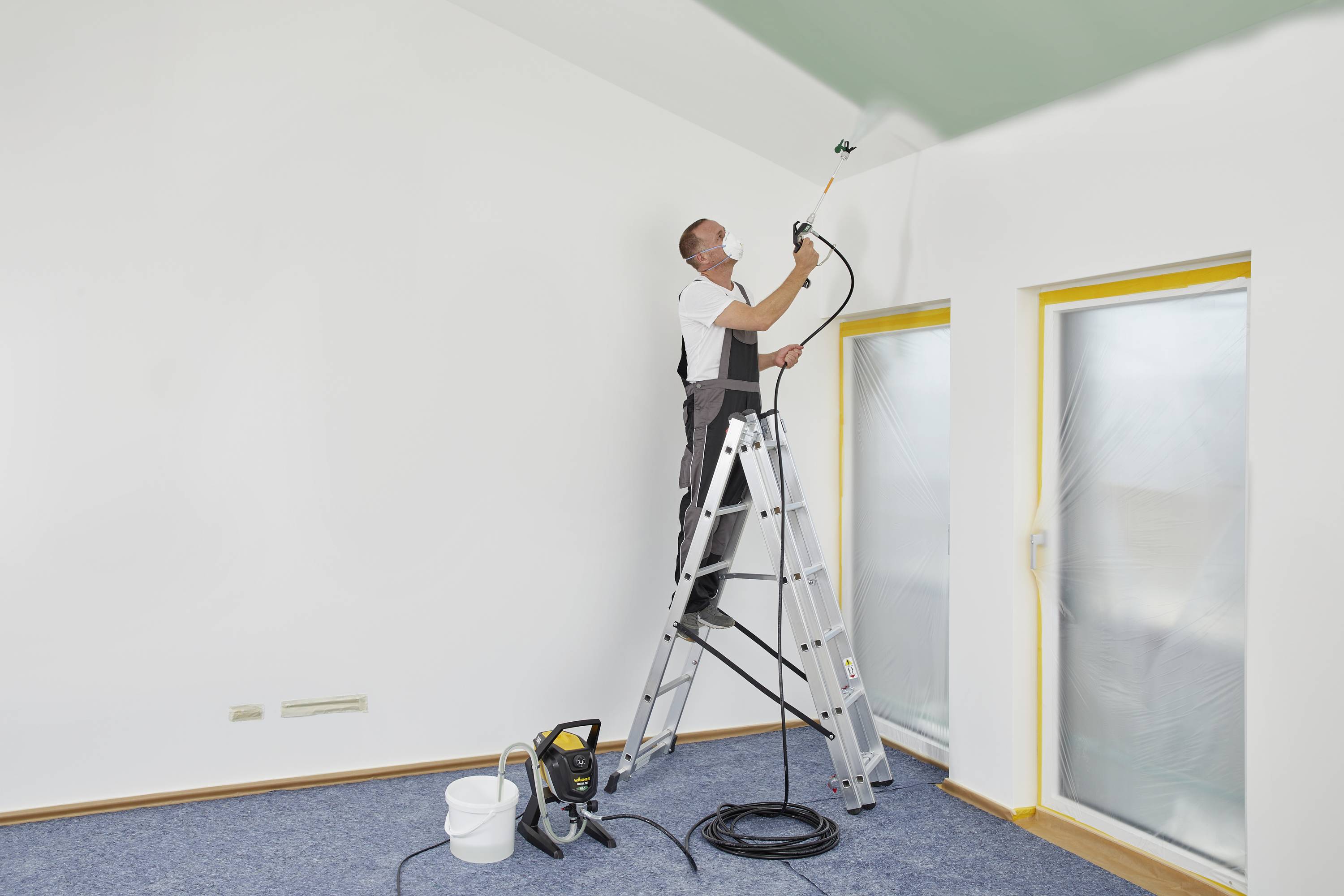 Ein Mann steht auf einer Leiter und streicht die Decke eines Zimmers mit grüner Farbe. Fensterscheiben sind mit Folie abgedeckt.