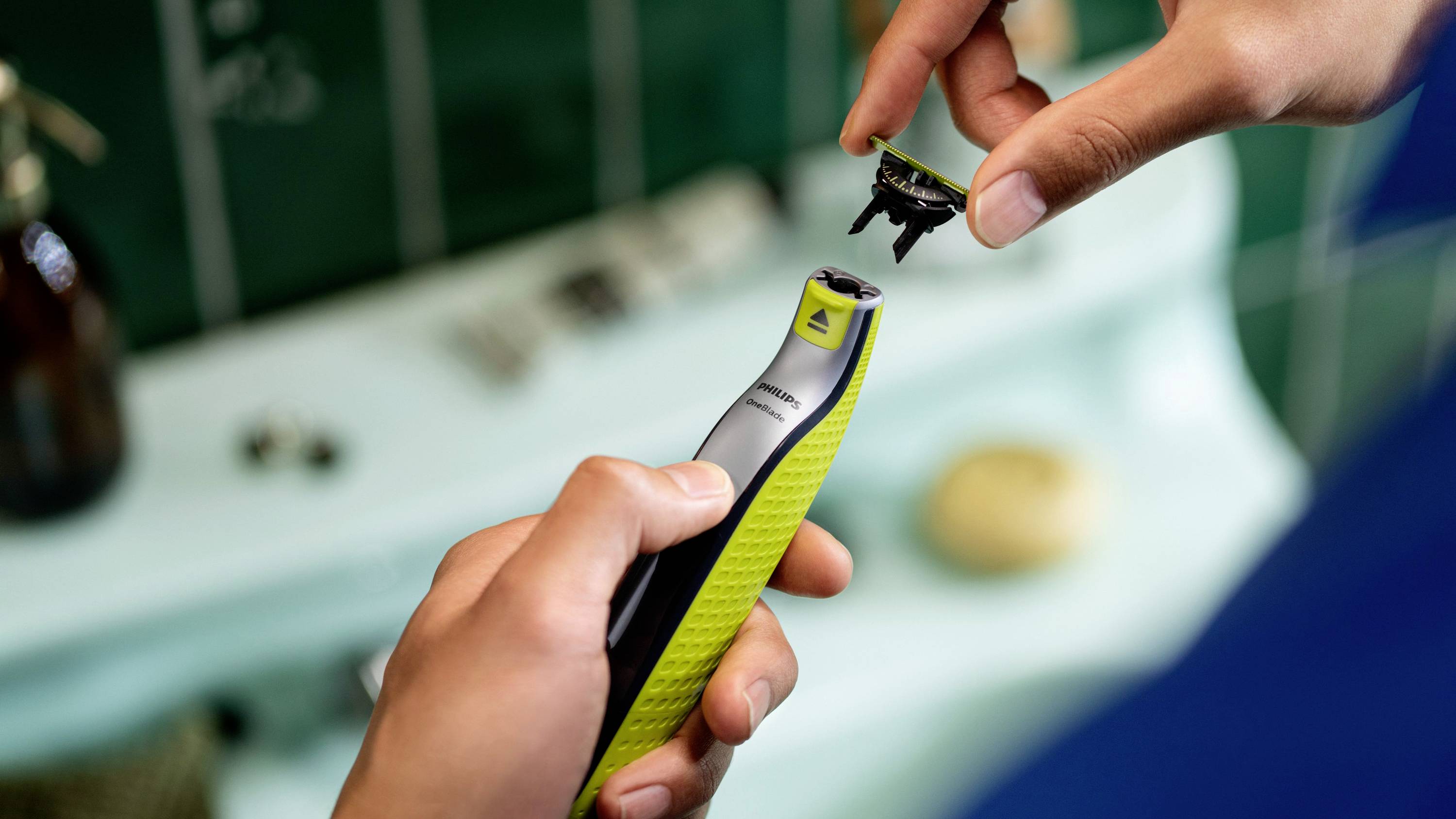 Eine Person hält einen elektrischen Rasierer und wechselt den Rasierkopf. Der Hintergrund zeigt verschwommene Badezimmerutensilien.