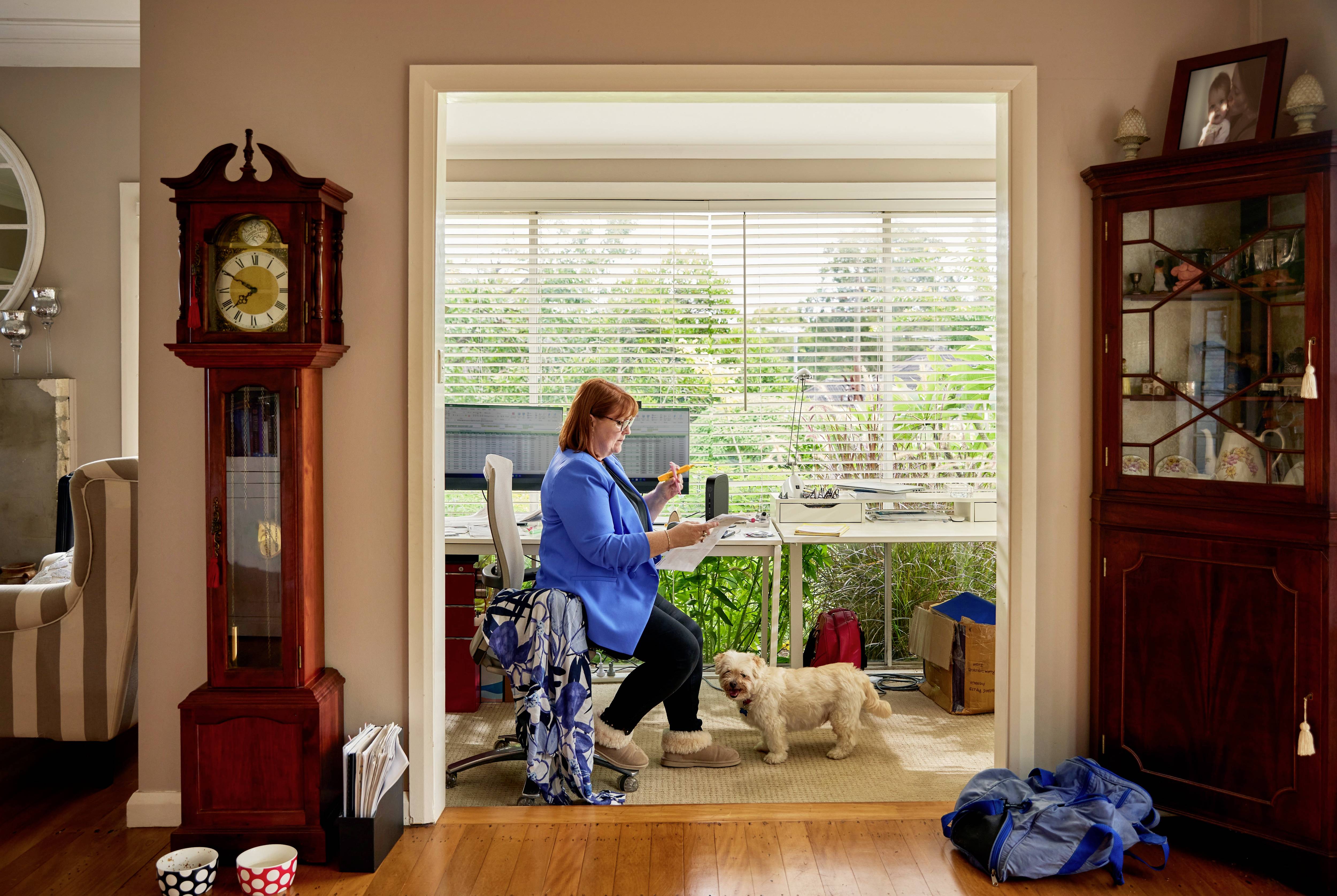 Eine Frau sitzt in ihrem Heimbüro und arbeitet an einem Schreibtisch. Ein kleiner Hund steht neben ihr. Fenster im Hintergrund bieten Blick ins Grüne.