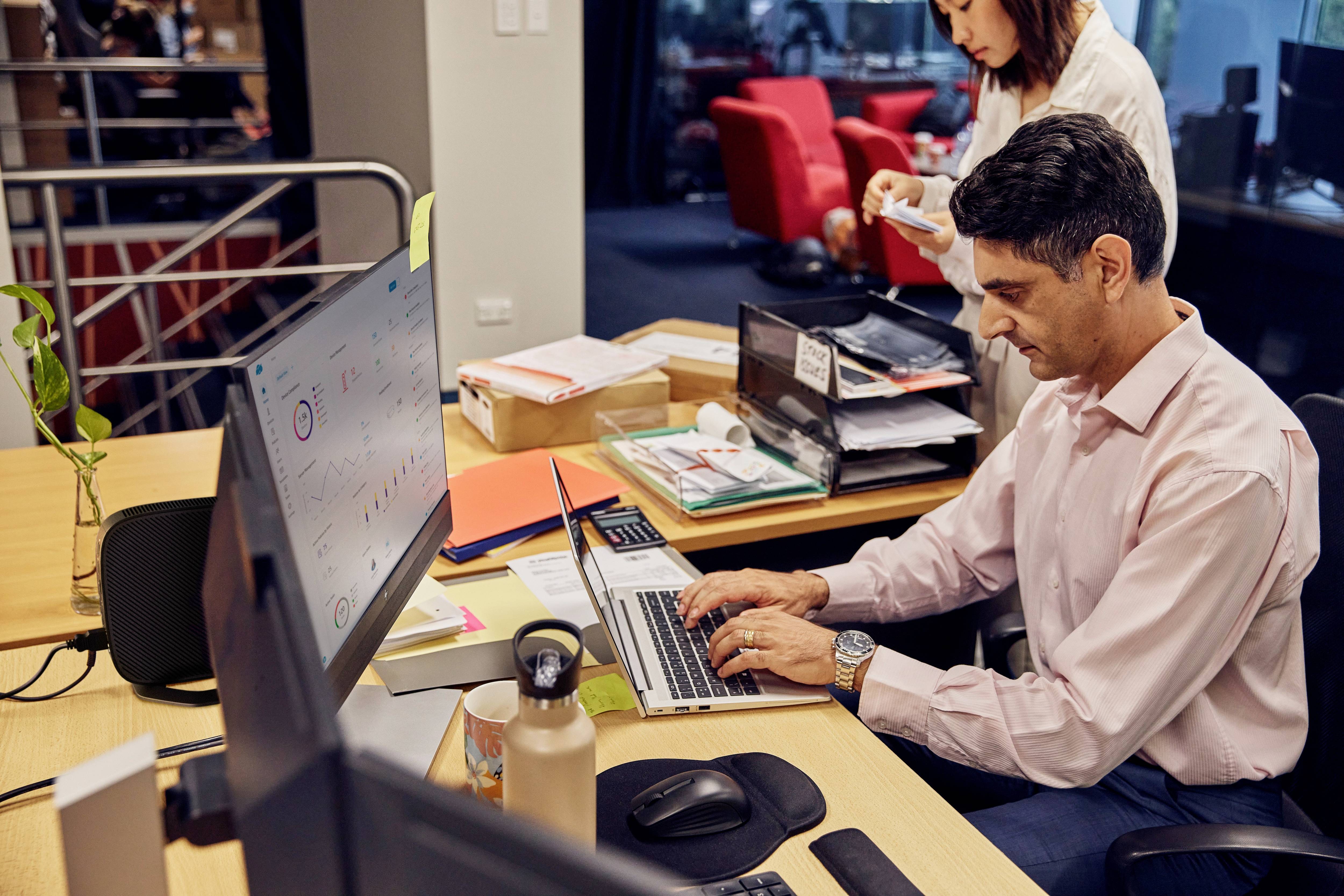 Ein Mann arbeitet an einem Laptop an einem Schreibtisch mit Monitoren. Eine Frau steht dahinter, hält Papiere. Büro im Hintergrund.