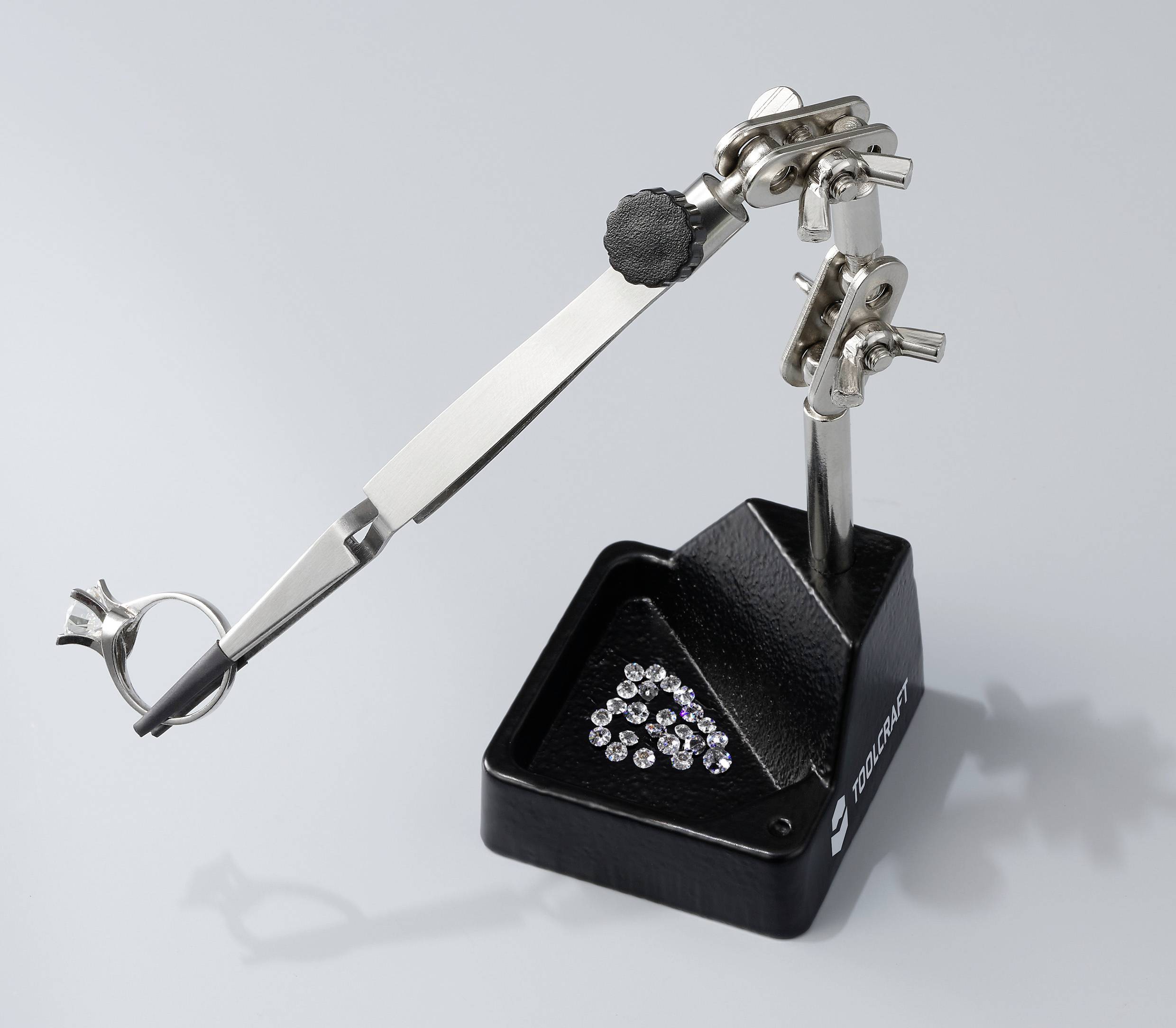 Ein Halterungswerkzeug mit Metallklammern und einer Lupe, verwendet für präzise Arbeiten wie Schmuckherstellung. Darunter liegen kleine Diamanten.