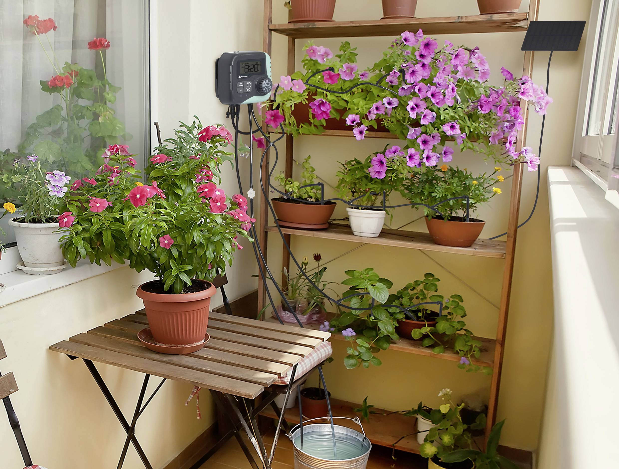 Ein Balkon mit Topfpflanzen auf einem Regal und Tisch, darunter bunte Blüten und ein Bewässerungseimer auf dem Boden.