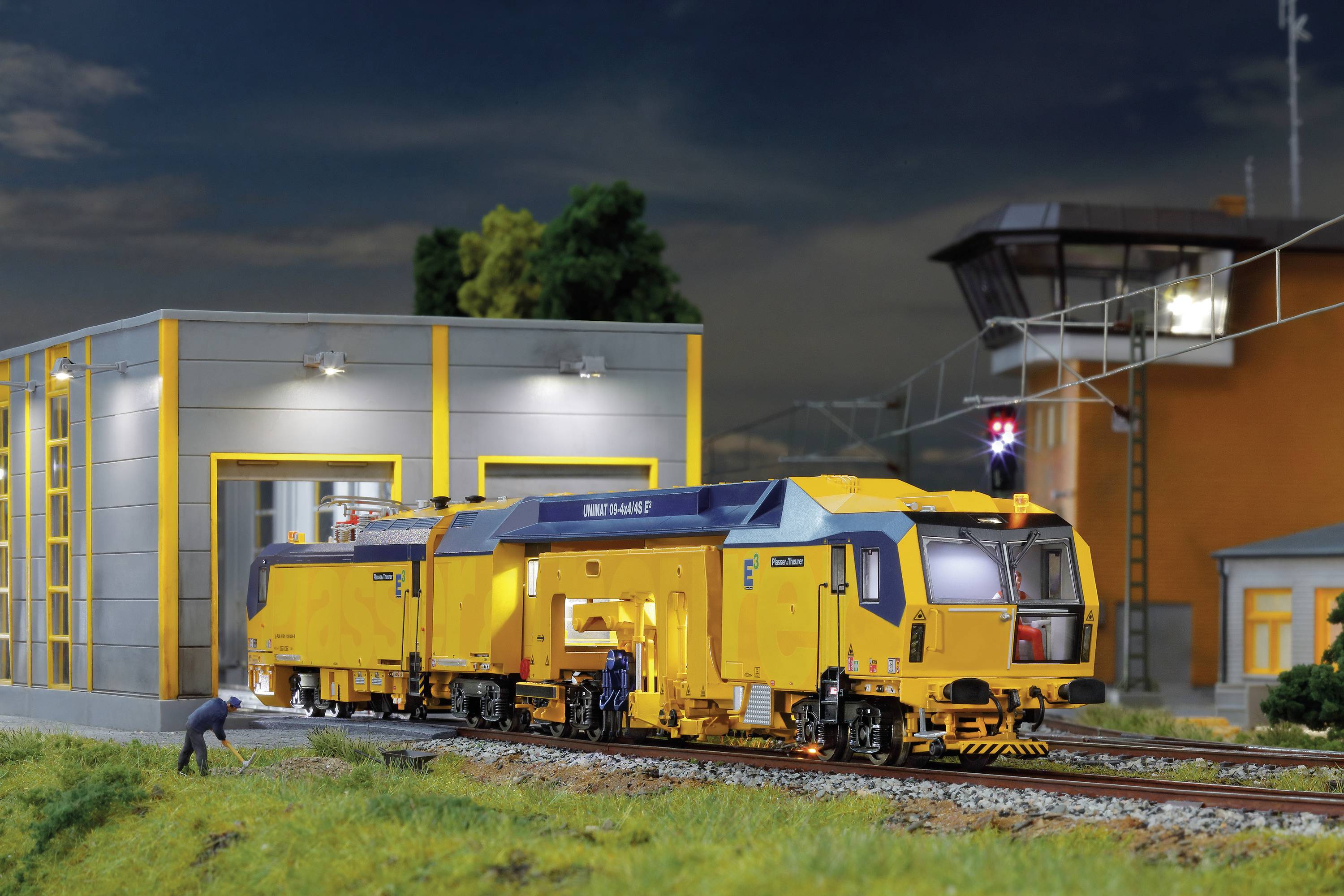 Eine gelbe Lokomotive steht auf einem Gleis vor einem beleuchteten Gebäude. Im Vordergrund arbeiten zwei Personen an der Lokomotive.