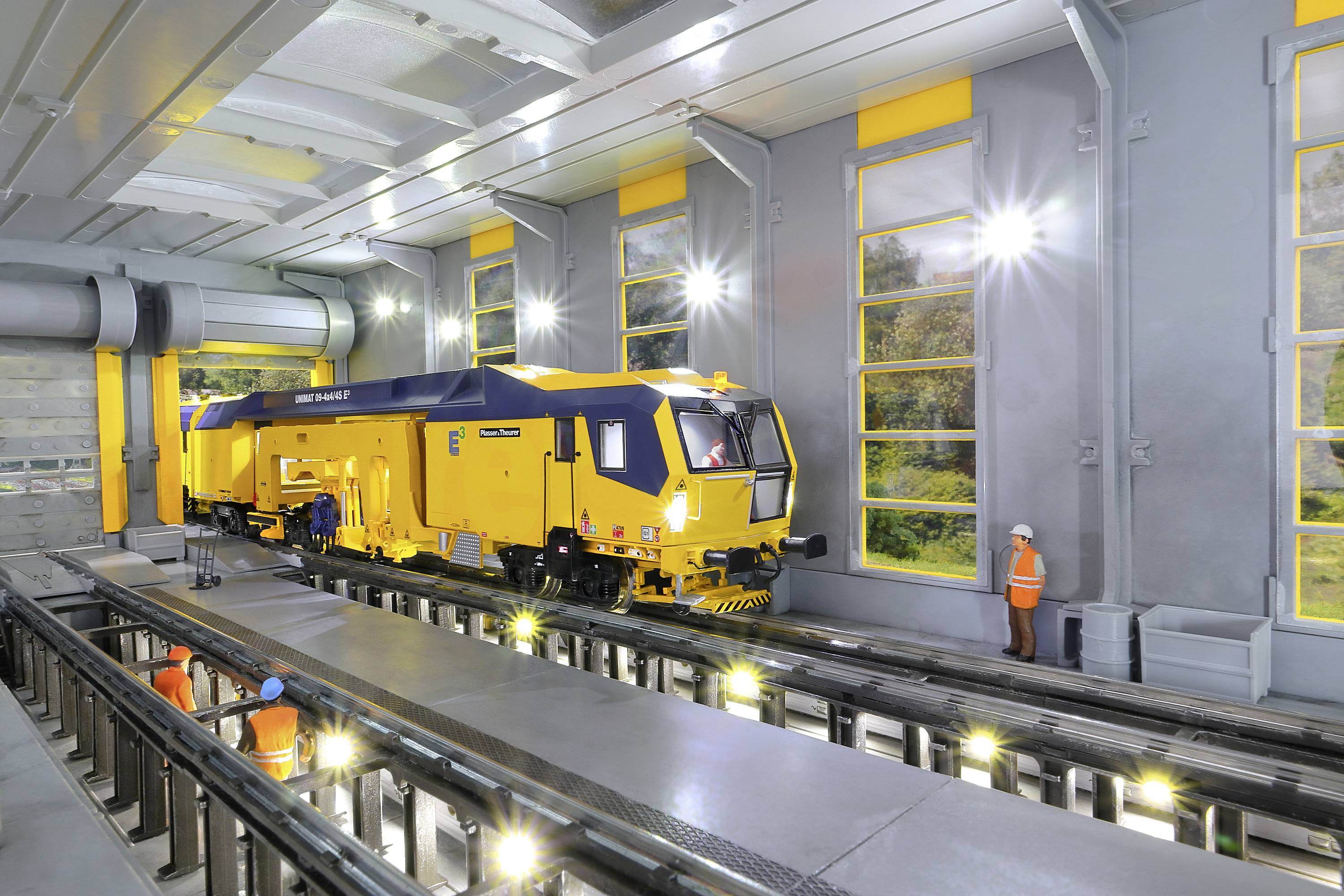Gelber Schienenfahrzeug-Roboter in Werkstatt mit erhöhten Gleisen und Arbeitern in orangefarbener Schutzkleidung.