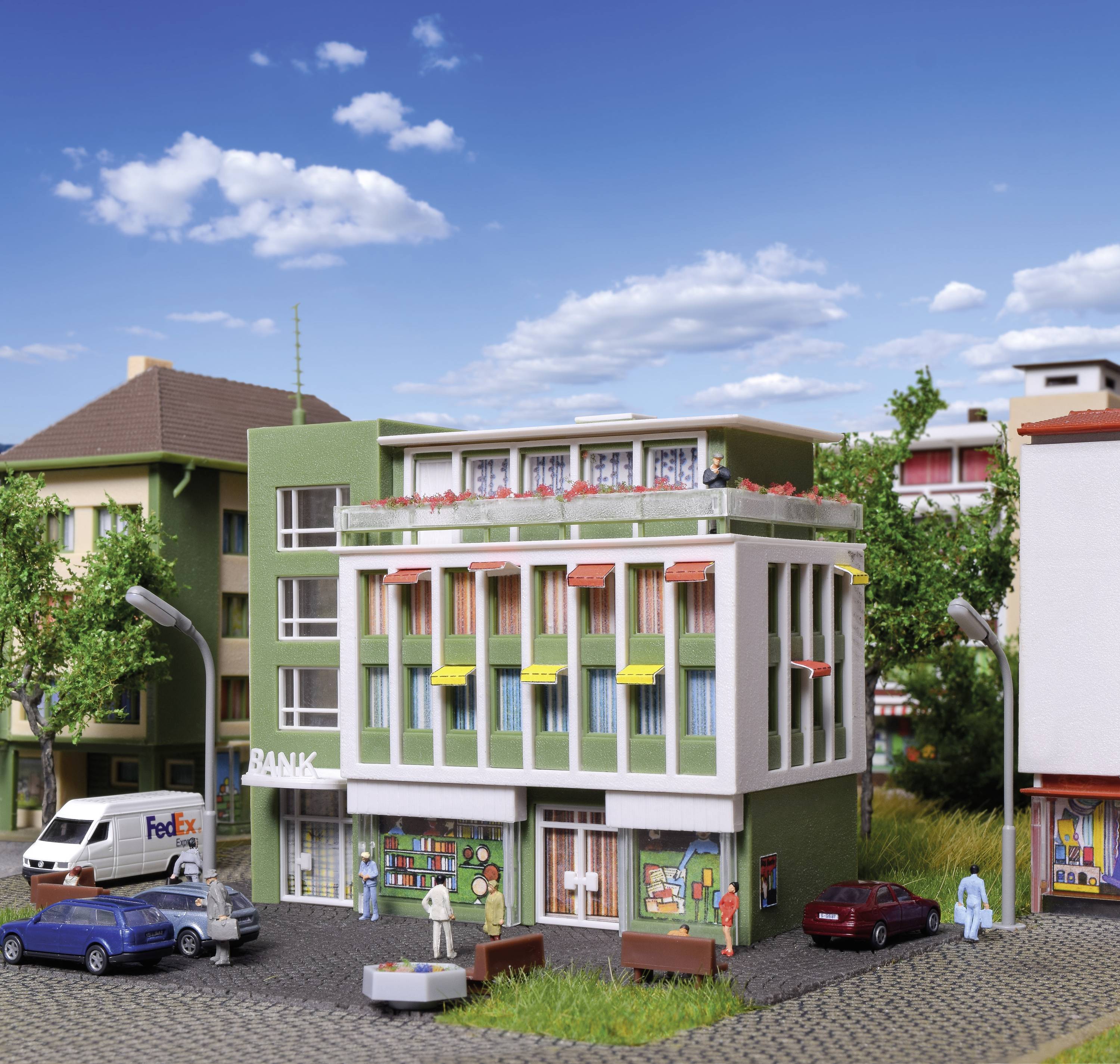 Mehrstöckiges Modellgebäude mit Geschäften im Erdgeschoss und Wohnungen darüber, umgeben von Modellfahrzeugen und Bäumen.