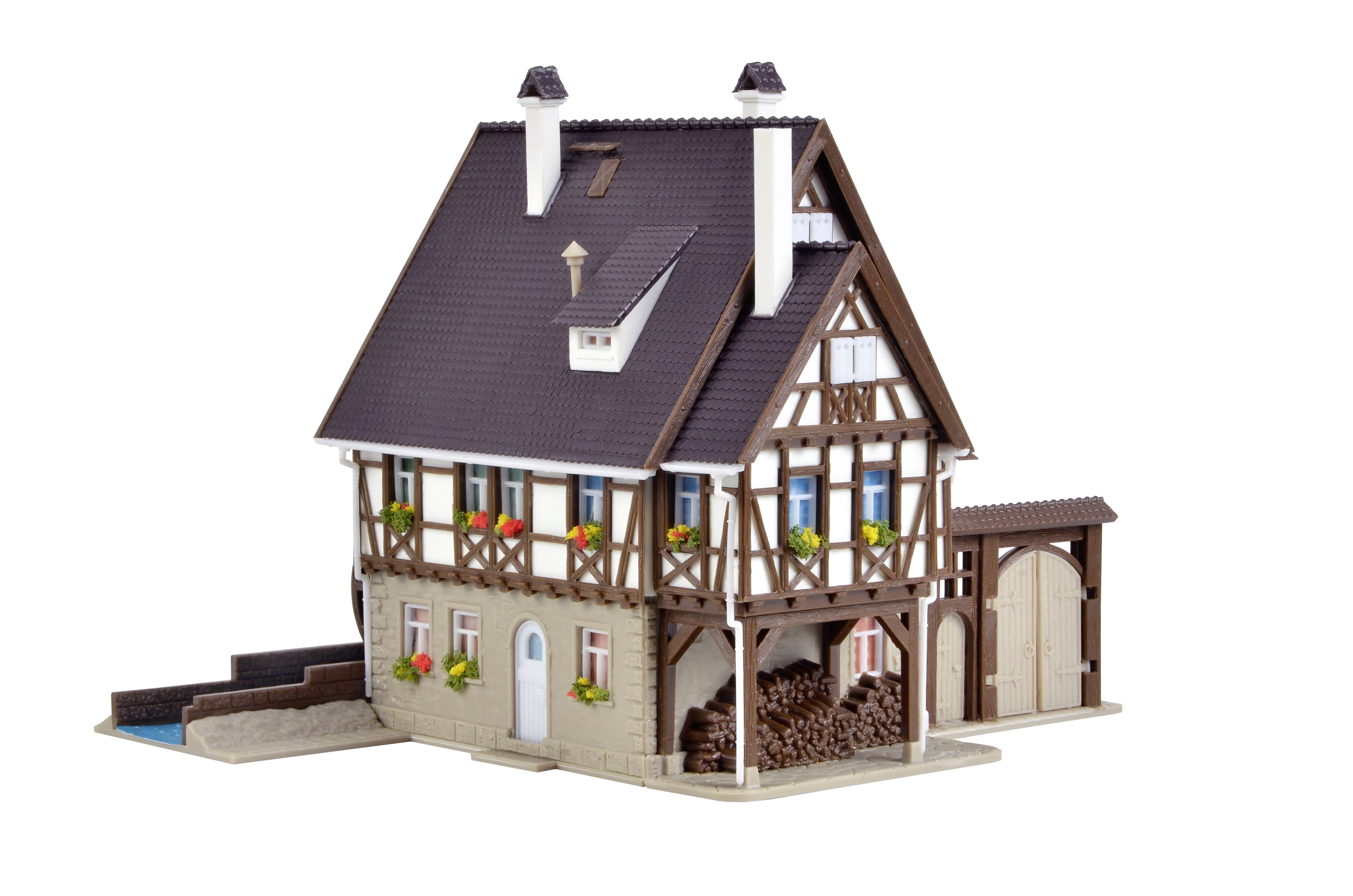 Ein Modell eines traditionellen Fachwerkhauses mit Holzbalken und Blumenschmuck an den Fenstern, neben einem Holzstapel.