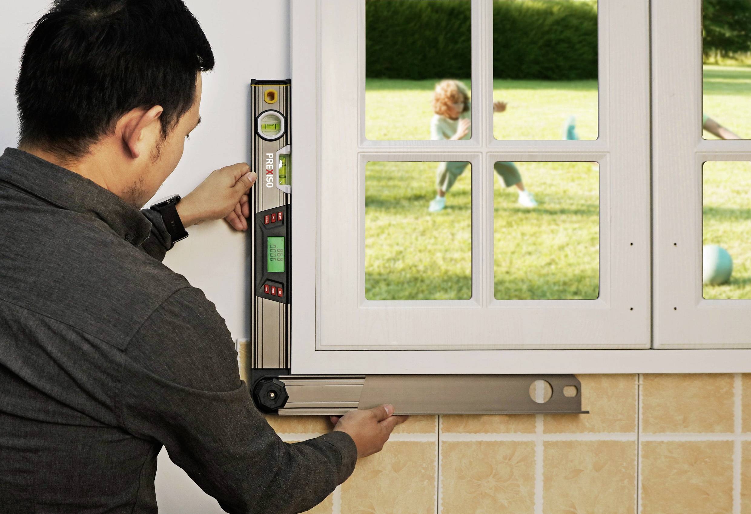 Ein Mann verwendet eine Wasserwaage, um ein Fenster auszurichten. Durch das Fenster ist ein Kind beim Spielen auf einer Wiese zu sehen.