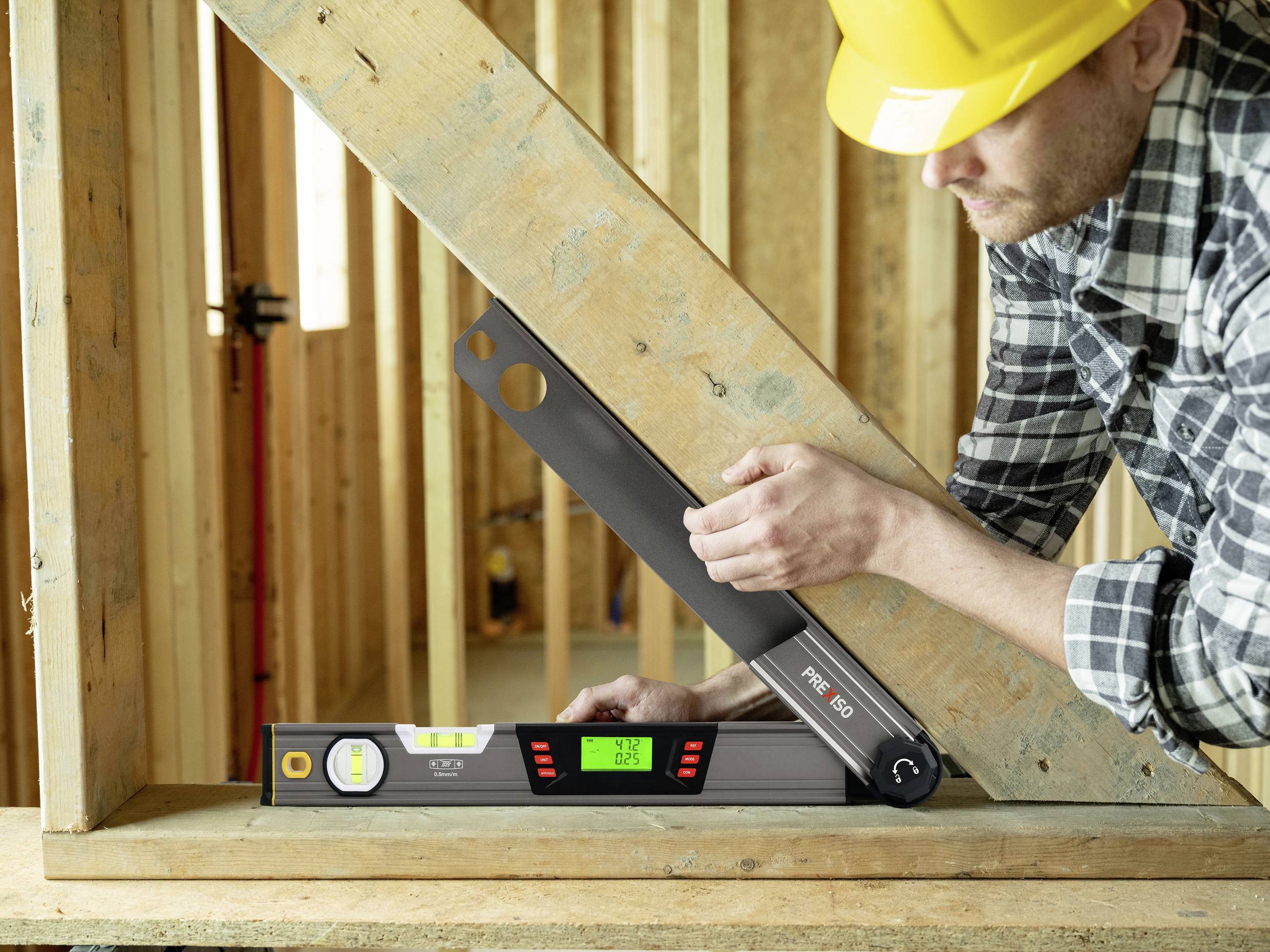 Ein Bauarbeiter in Schutzhelm verwendet ein digitales Winkelmessgerät, um den Winkel eines Holzrahmens in einem Rohbau zu überprüfen.