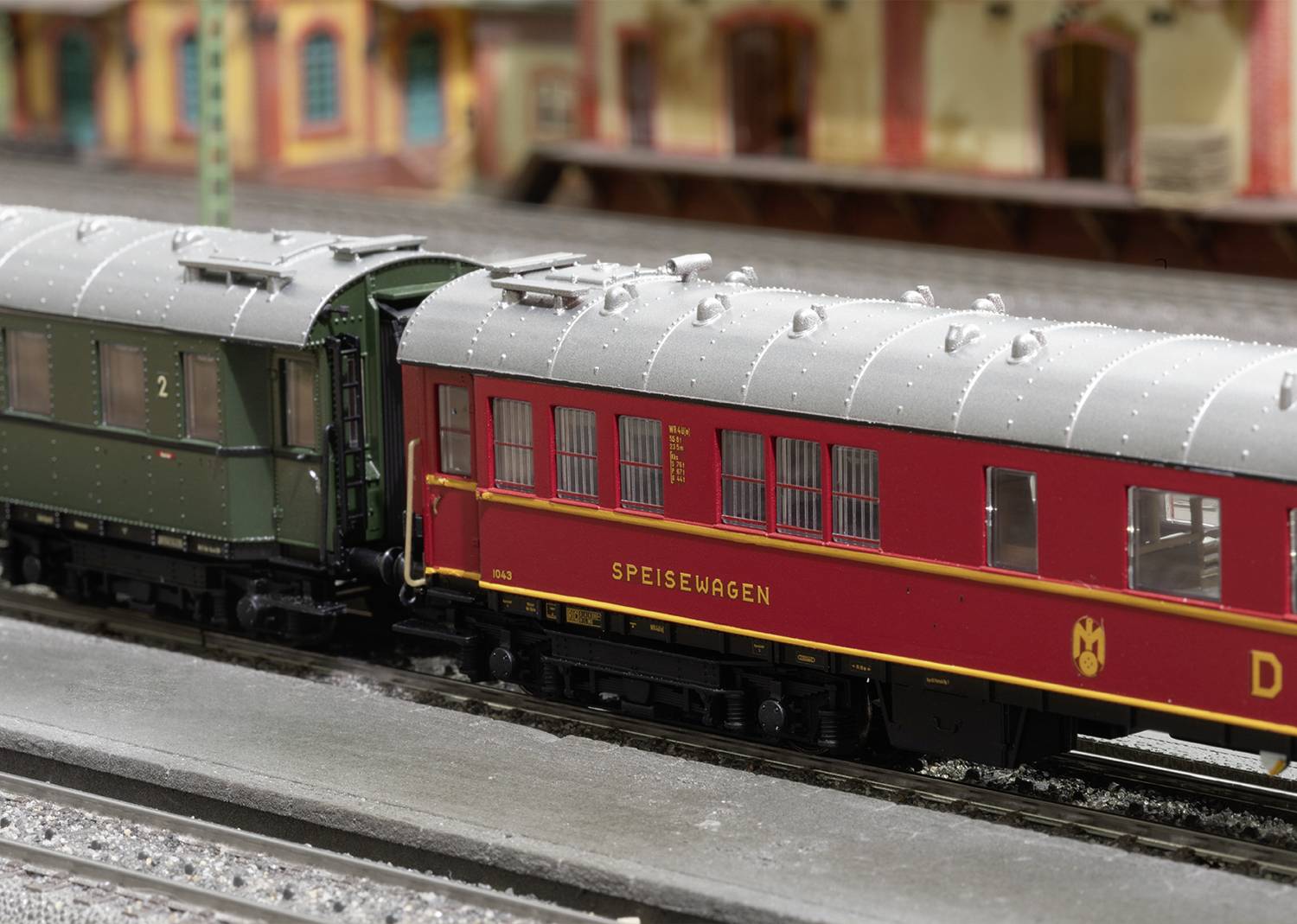 Ein Modellzug mit einem roten Speisewagen und einem grünen Personenwagen steht auf einem Gleis vor einem historischen Bahnhofsgebäude.