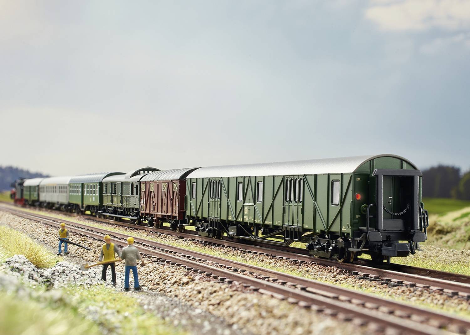 Eine Modellbahnstrecke mit mehreren Güterwaggons. Am Bahndamm stehen Figuren in Arbeitskleidung und führen Arbeiten durch.