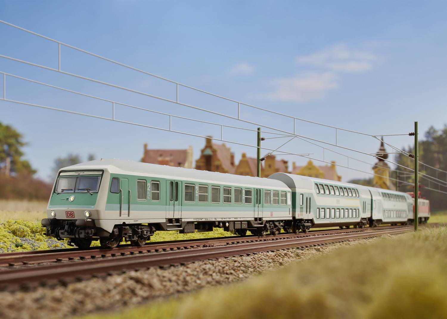 Ein Modellzug im Maßstab fährt auf Gleisen durch eine ländliche Szenerie mit Bäumen und einem Gebäude im Hintergrund unter klarem Himmel.