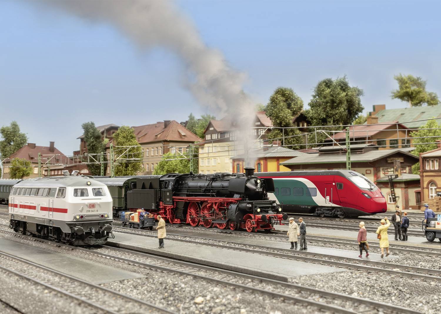 Eisenbahnmodellanlage mit mehreren Zügen: Dampflok mit Rauch und moderner Zug im Hintergrund. Menschen auf dem Bahnsteig.