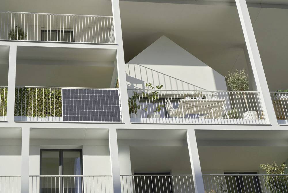Mehrstöckiges modernes Gebäude mit Balkonen. Ein Balkon zeigt Solarpanel, Pflanzen und Gartenmöbel. Sonne beleuchtet die Fassade.