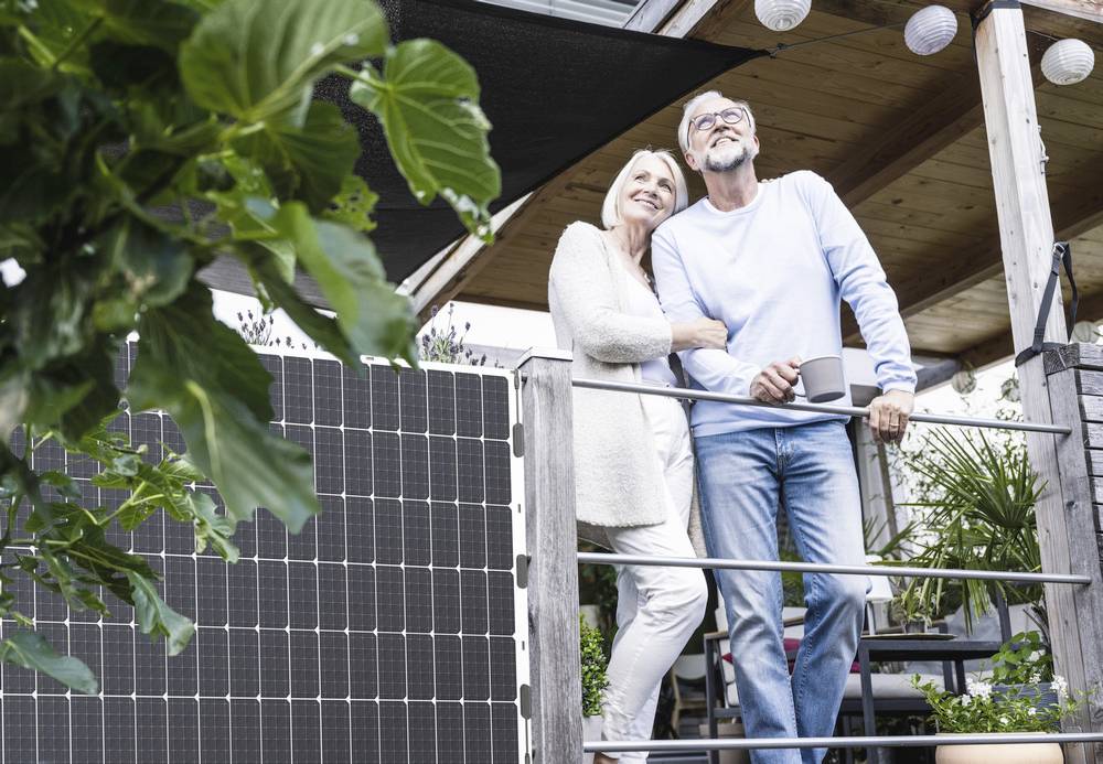 Ein älteres Paar steht auf einer Terrasse neben Solarpaneelen. Sie schauen gemeinsam in die Ferne und wirken zufrieden und entspannt.