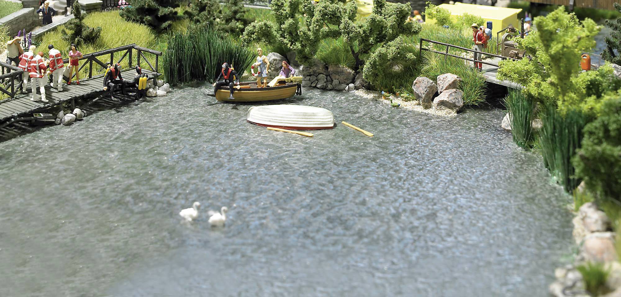 Ein Teich mit Booten, umgeben von Bäumen und Gräsern. Menschen stehen auf Holzbrücken und blicken auf das Wasser. Zwei Enten schwimmen.
