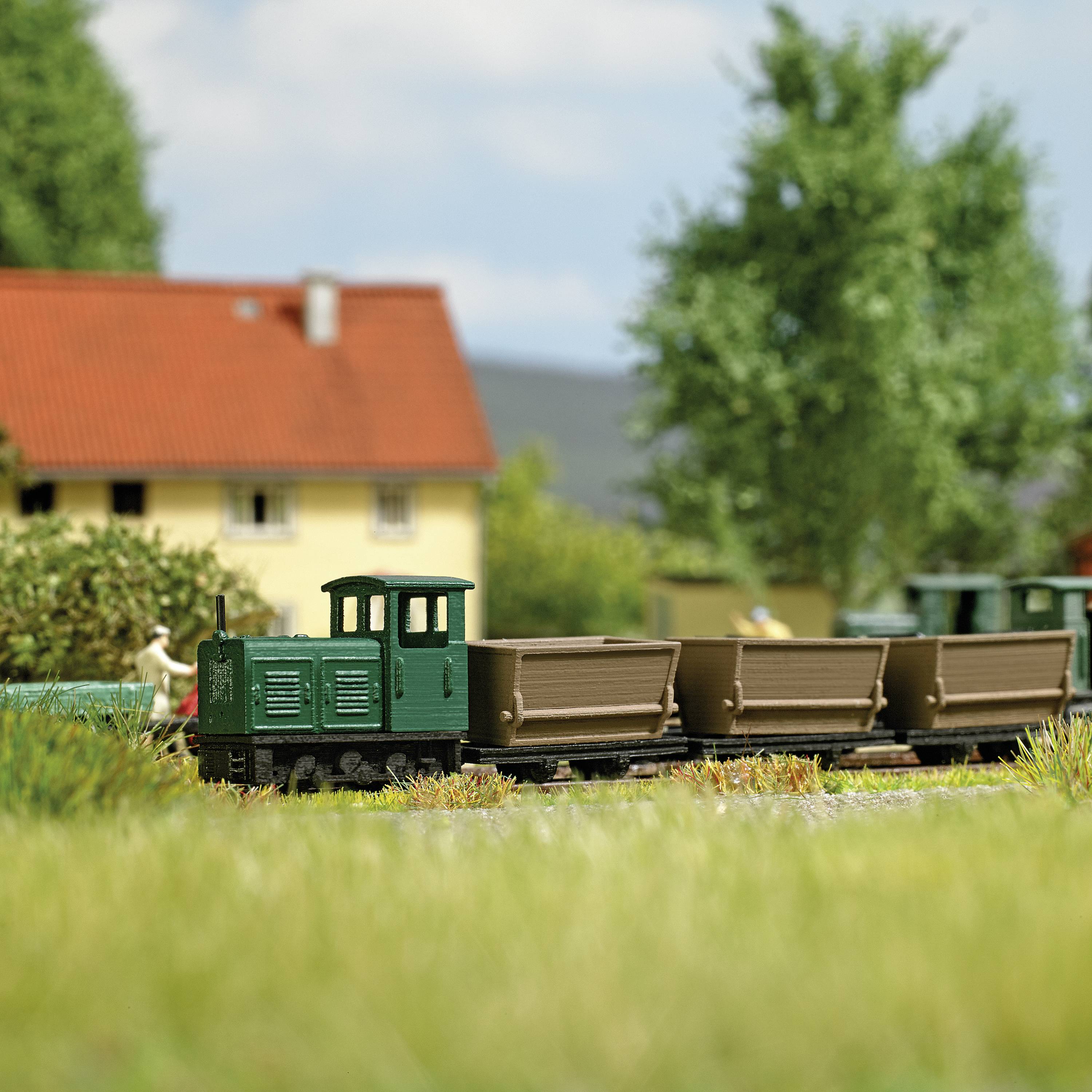 Eine modellhafte Szenerie zeigt eine kleine, grüne Lokomotive vor gelben Gebäuden und Bäumen, die Ruhe und Ländlichkeit vermitteln.