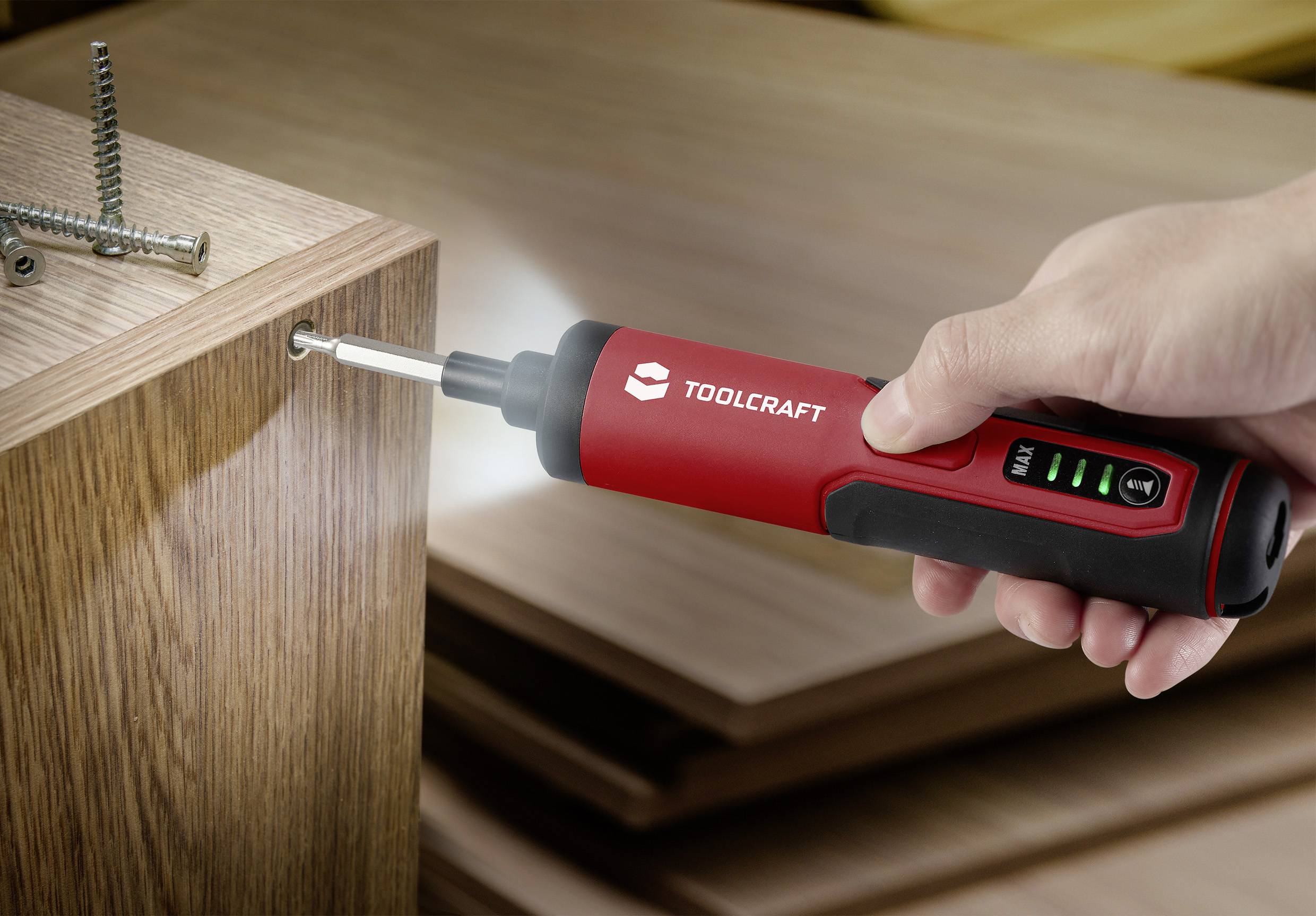 Eine Hand hält einen roten elektrischen Schraubendreher, der eine Schraube in ein Holzbrett schraubt. Drei Schrauben sind im Brett befestigt.