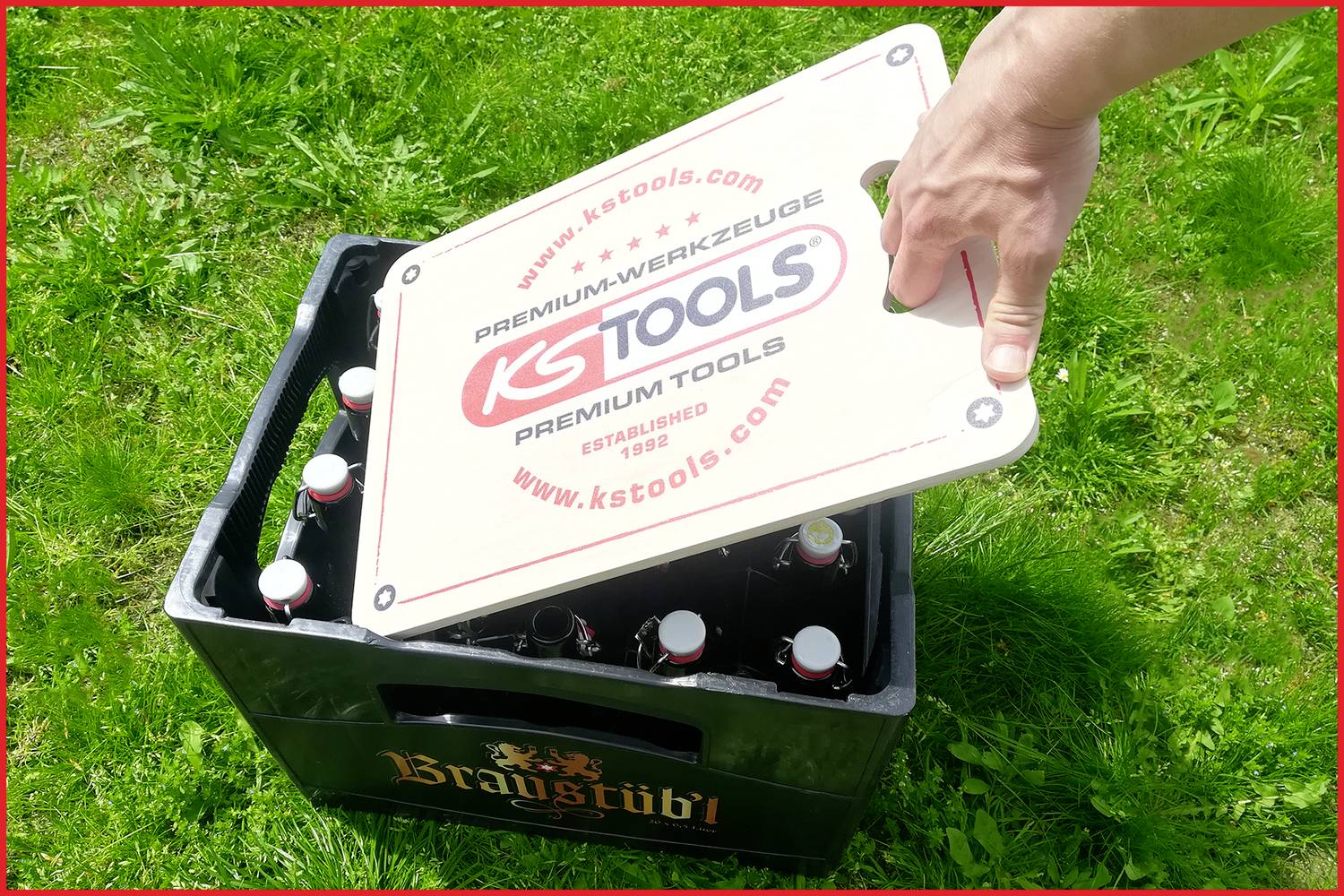 Eine Hand hebt einen bedruckten Kastenöffner von einer Getränkekiste auf einer Wiese. Auf dem Öffner steht 'KS Tools'.