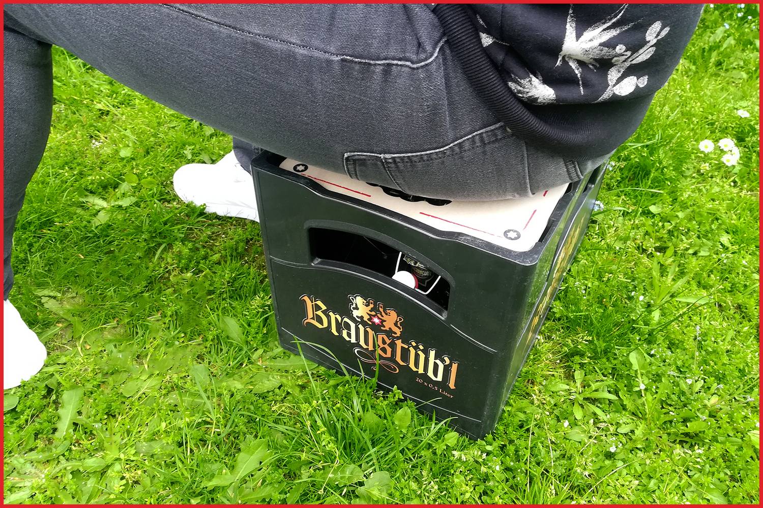 Person sitzt auf einer leeren Getränke-Kiste der Marke BrauStübl auf einer Wiese.