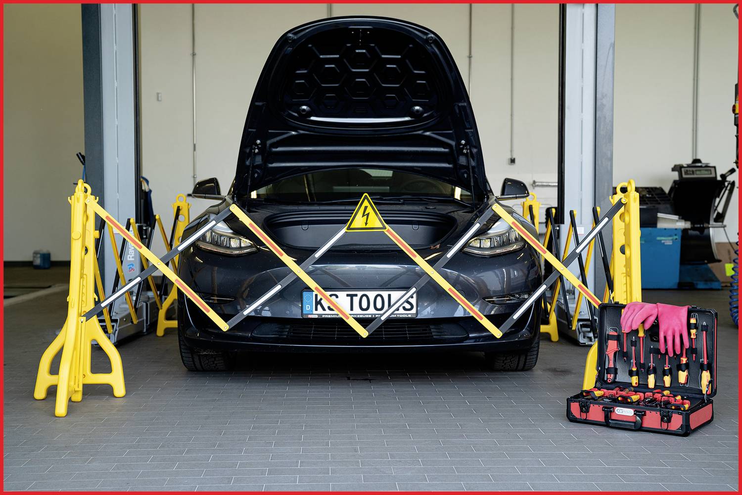 Ein Auto mit geöffneter Motorhaube steht in einer Werkstatt. Warnbänder und Warnschild deuten auf elektrische Wartungsarbeiten hin.
