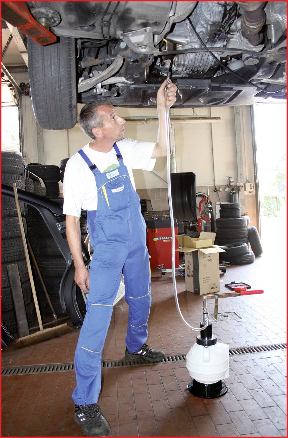 Mechaniker in blauer Arbeitskleidung arbeitet unter einem angehobenen Auto in einer Werkstatt. Er benutzt eine Pumpe zur Flüssigkeitsentnahme. Im Hintergrund sind Reifen und Werkzeuge zu sehen.