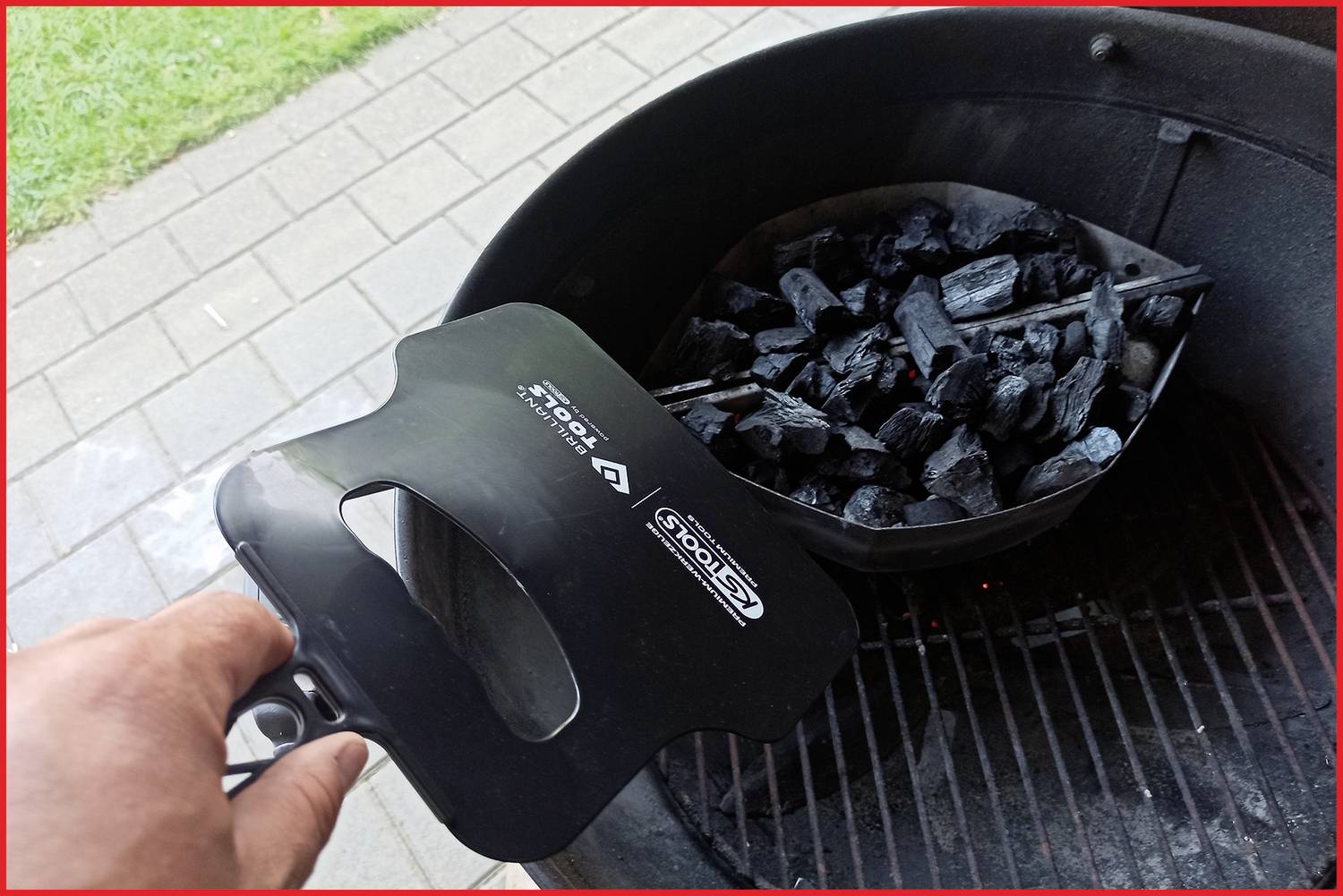 Eine Hand hält einen Anzündkamin über einem Grill mit glühender Holzkohle. Der Hintergrund zeigt eine gepflasterte Terrasse.