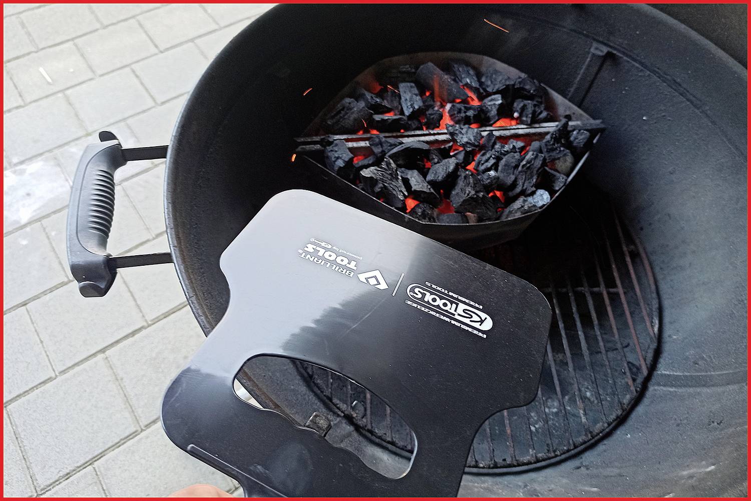 Ein Grill mit glühender Kohle in einem Kohlekorb. Eine Grillabdeckung wird am Rand des Grills gehalten, vermutlich zum Schließen.