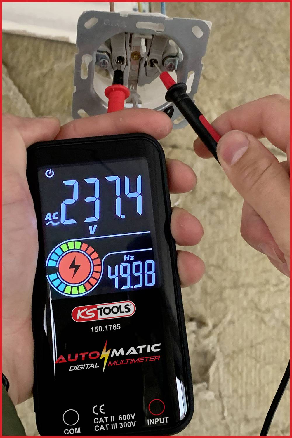 Eine Hand hält ein digitales Multimeter, das 237,4 AC Volt anzeigt, während es mit einer Steckdose verbunden ist.