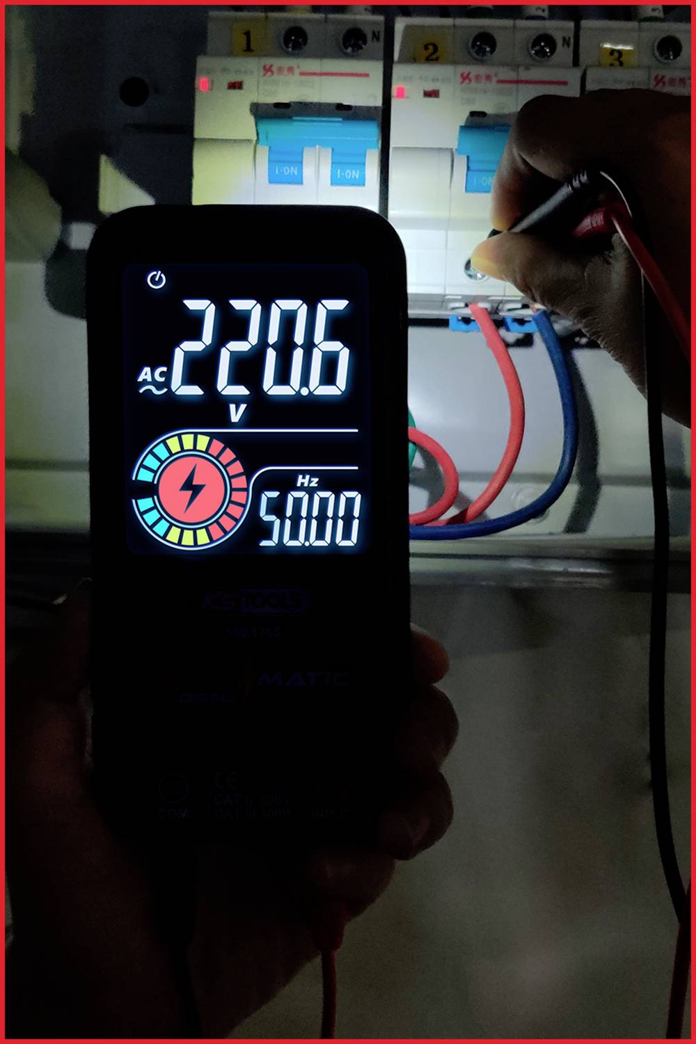 Eine Person misst mit einem digitalen Spannungsmessgerät 220,6 V AC bei 50,0 Hz an einem offenen Sicherungskasten mit angeschlossenen Drähten.
