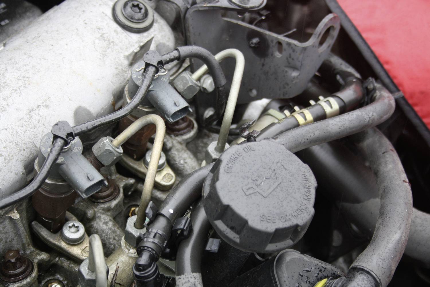Ein Motorraum mit verschiedenen Schläuchen, Kabeln und Metallteilen. Im Vordergrund ist ein runder Ölverschlussdeckel zu sehen.