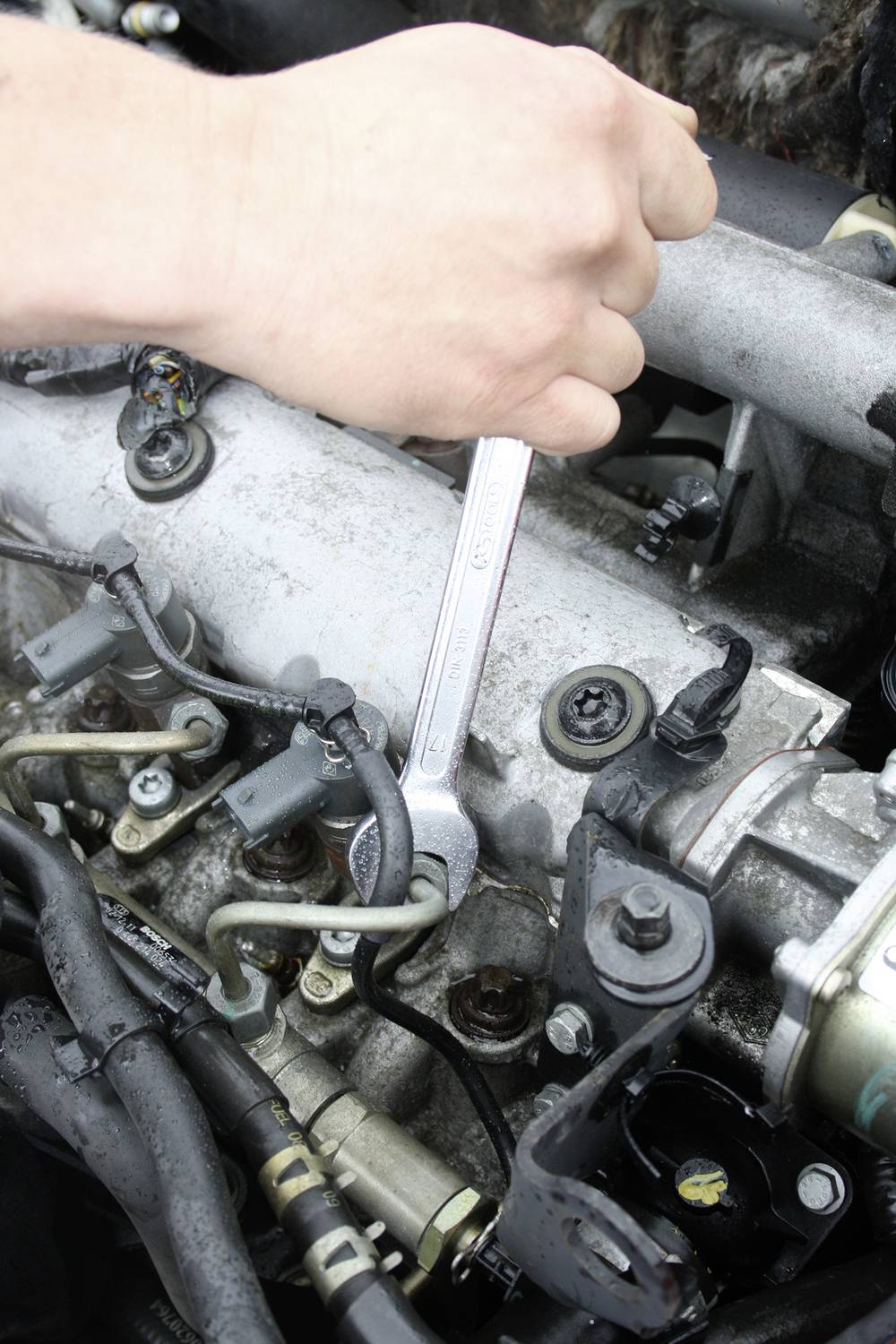 Eine Hand hält einen Schraubenschlüssel, der an einem Motor arbeitet. Der Motor zeigt mehrere Rohre und Kabel.