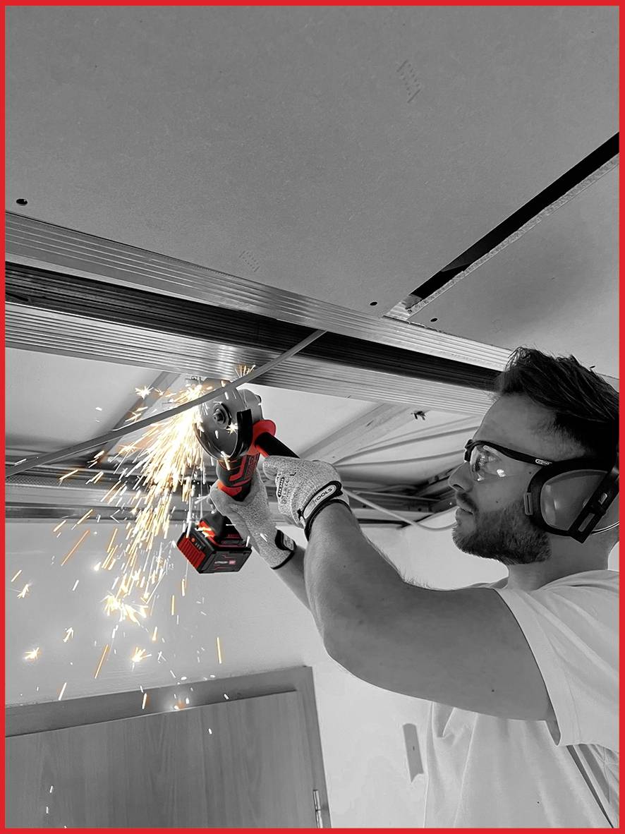 Ein Mann mit Schutzbrille und Gehörschutz schneidet Metallstreben an der Decke mit einem Winkelschleifer und es sprühen Funken.