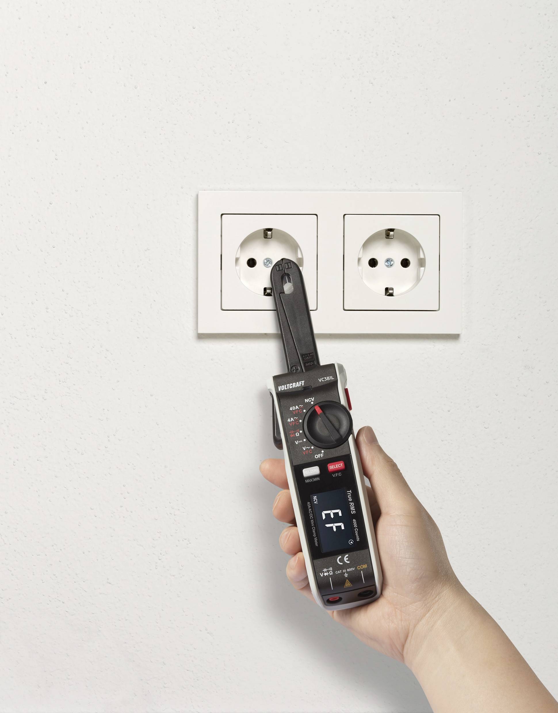 Eine Hand hält ein Multimeter mit einer Sonde an eine Doppelsteckdose an der Wand, das Display zeigt Messwerte.