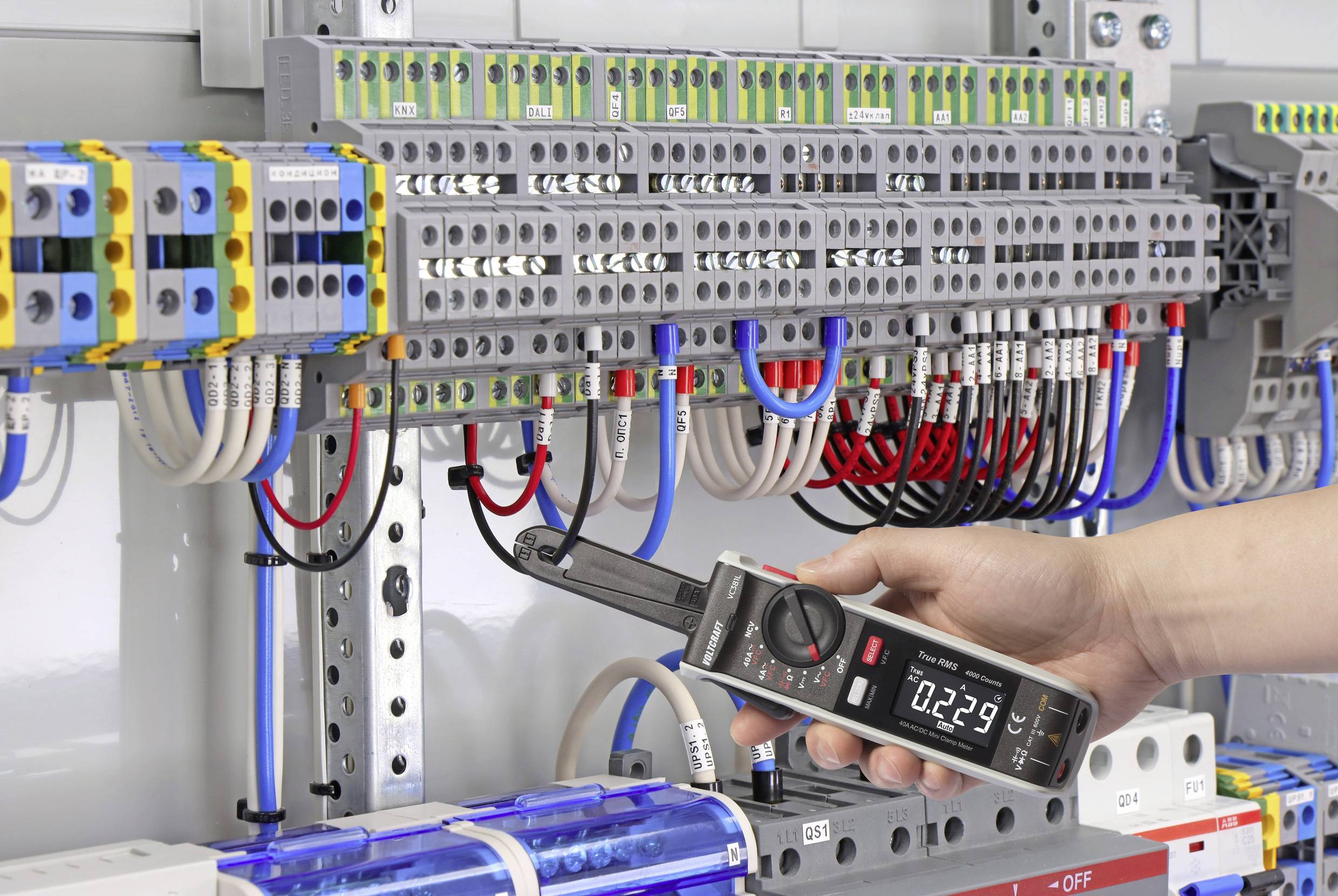 Eine Hand hält ein Multimeter, um den Strom in einem Schaltschrank mit vielen Kabeln und Schaltern zu messen. Das Display zeigt '0.229'.