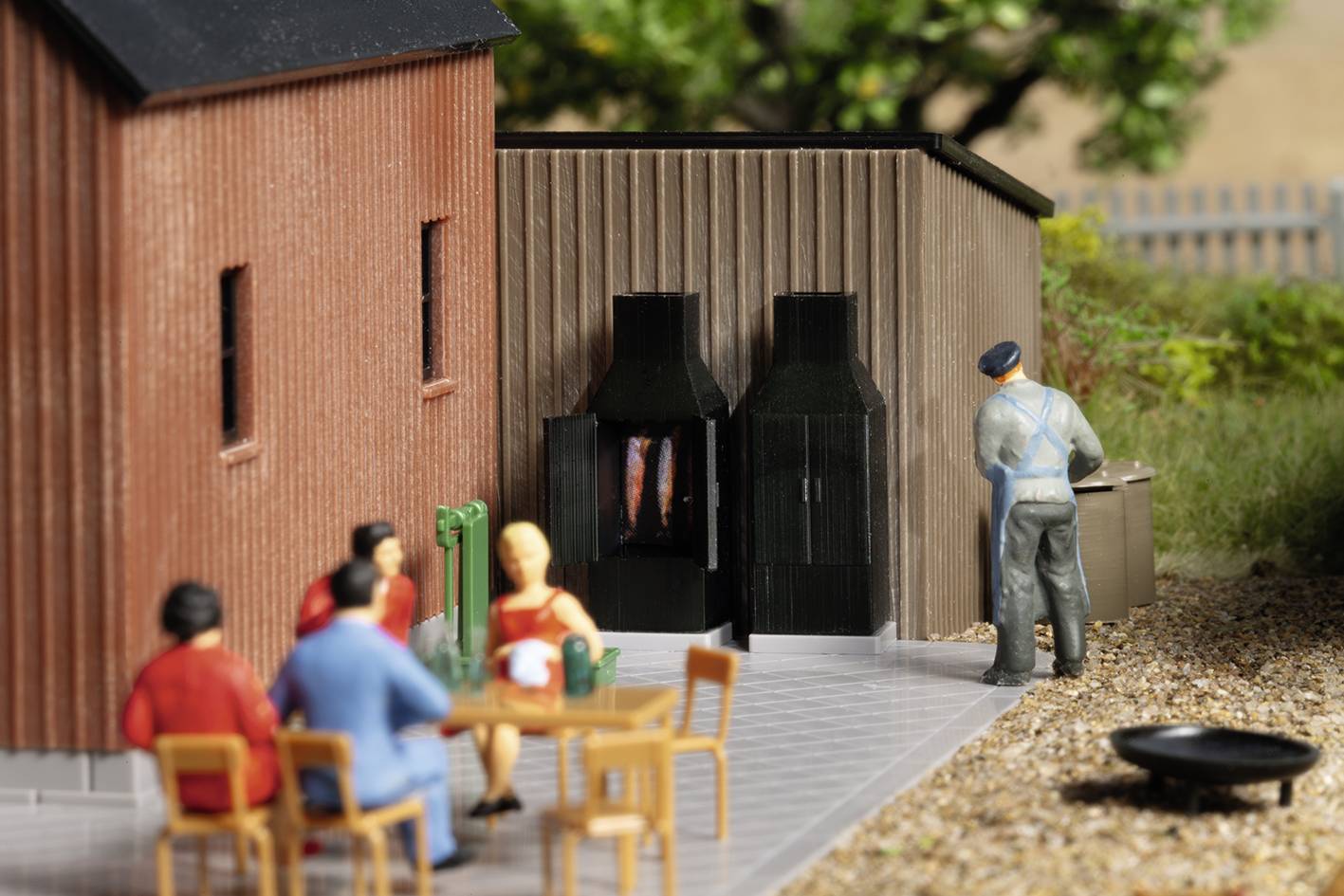 Modelleisenbahn-Diorama: Fünf Miniaturfiguren draußen; drei sitzen an einem Tisch, einer steht vor einem Grill, Holzgebäude im Hintergrund.