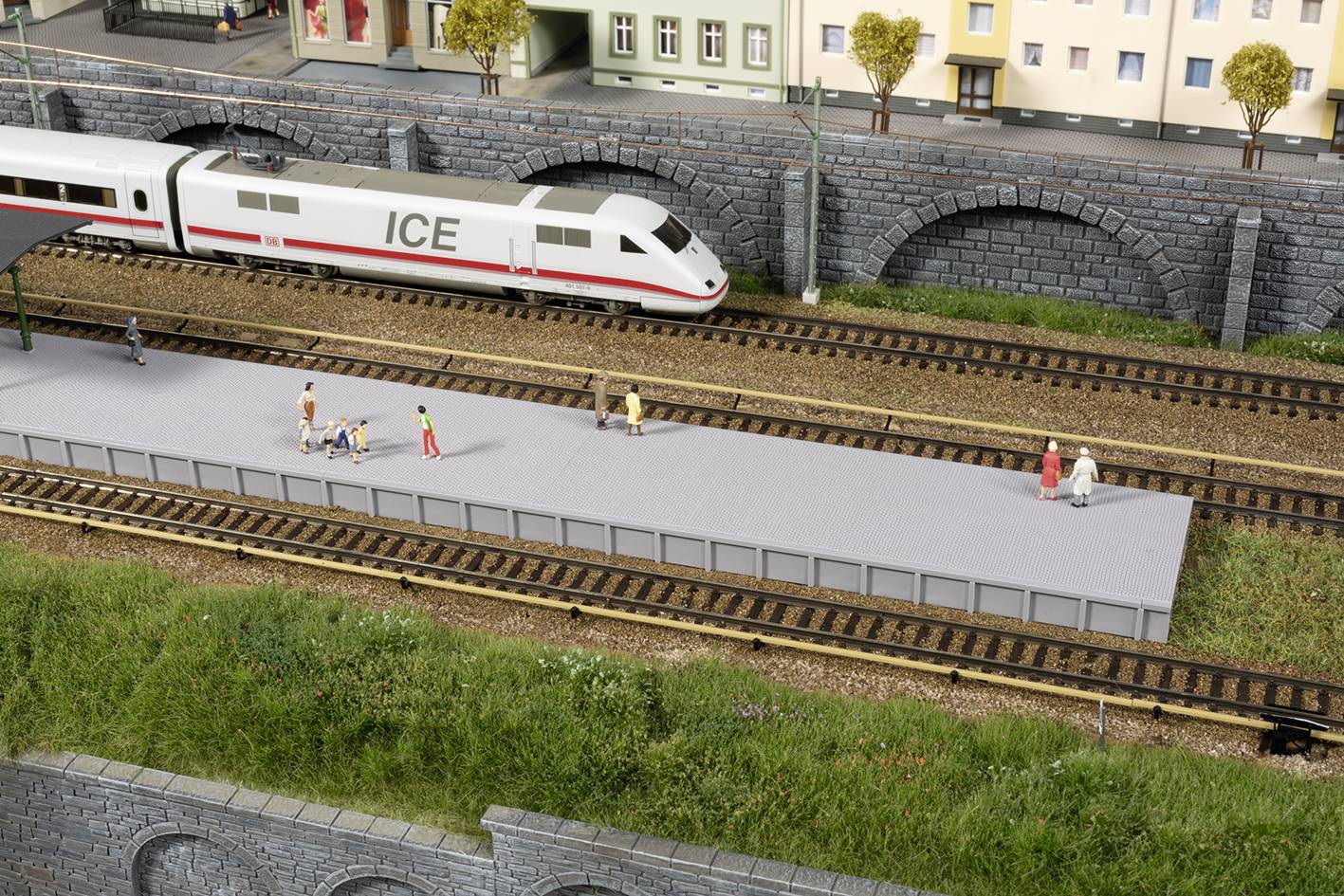 Ein ICE-Zug hält an einem Modellbahnhof mit mehreren Figuren auf dem Bahnsteig. Im Hintergrund sind Gebäude und Brücken zu sehen.