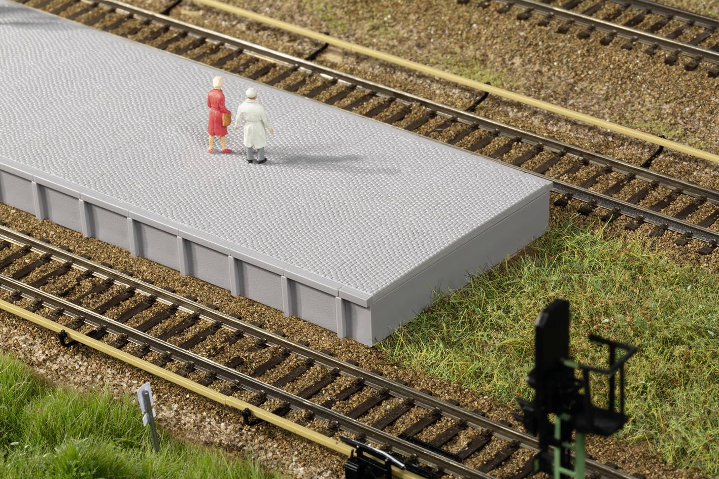 Zwei Figuren stehen auf einem Modellbahnsteig, umgeben von Schienen und Gras. Eine Signalampel ist am unteren Bildrand sichtbar.