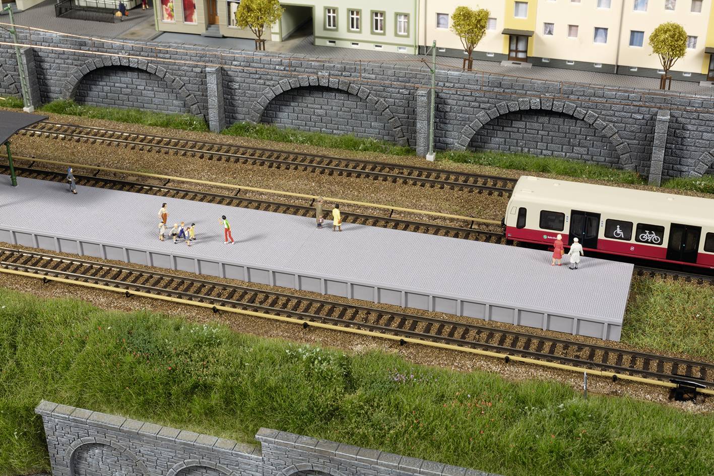 Ein Modellbahnsteig mit wartenden Figuren, daneben ein Zug. Hinten ist eine Häuserreihe und eine Steinbrücke zu sehen.