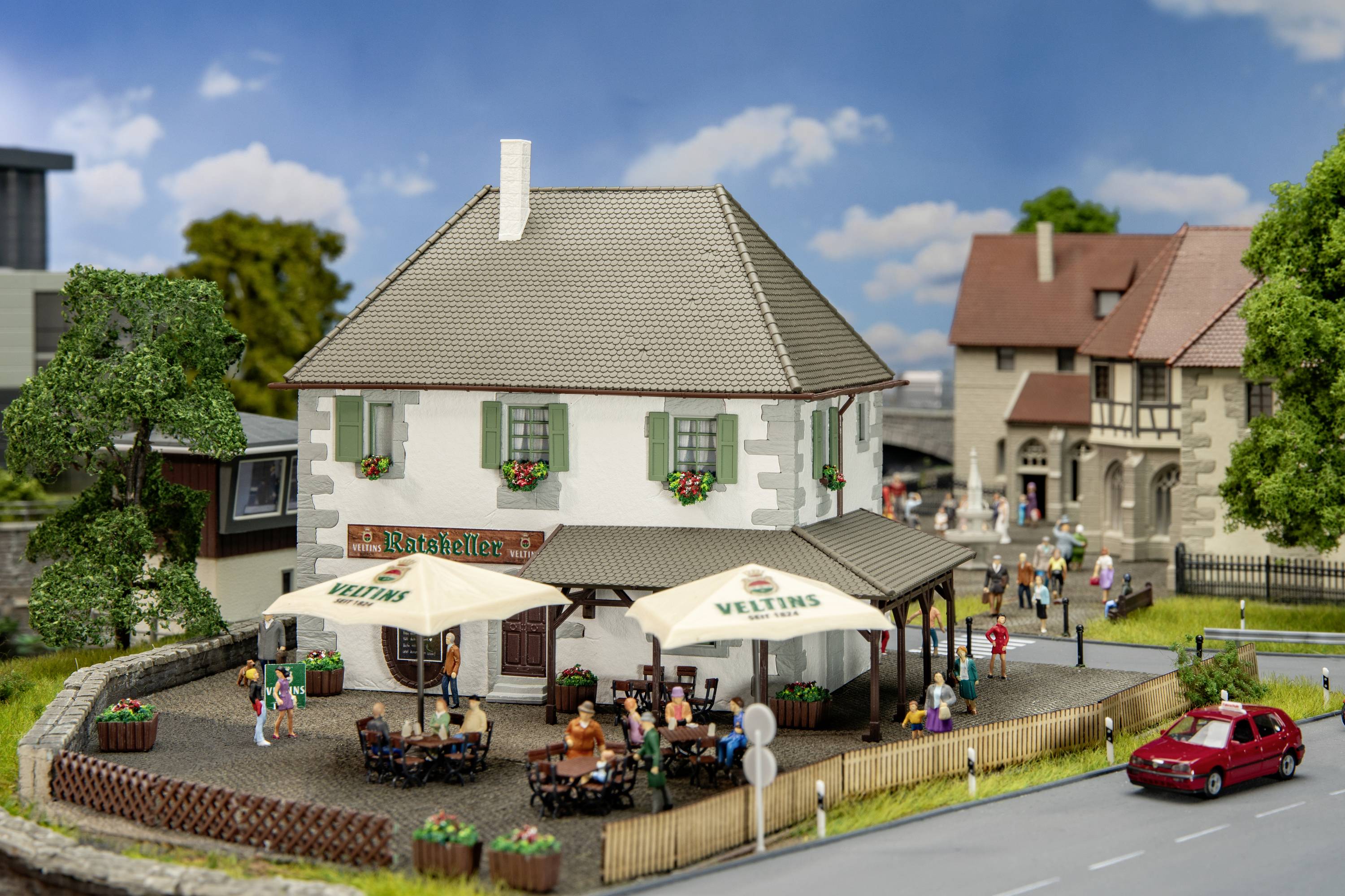 Ein Modell einer deutschen Dorfkneipe mit Terrasse und Menschen unter Sonnenschirmen. Häuser und Bäume im Hintergrund.