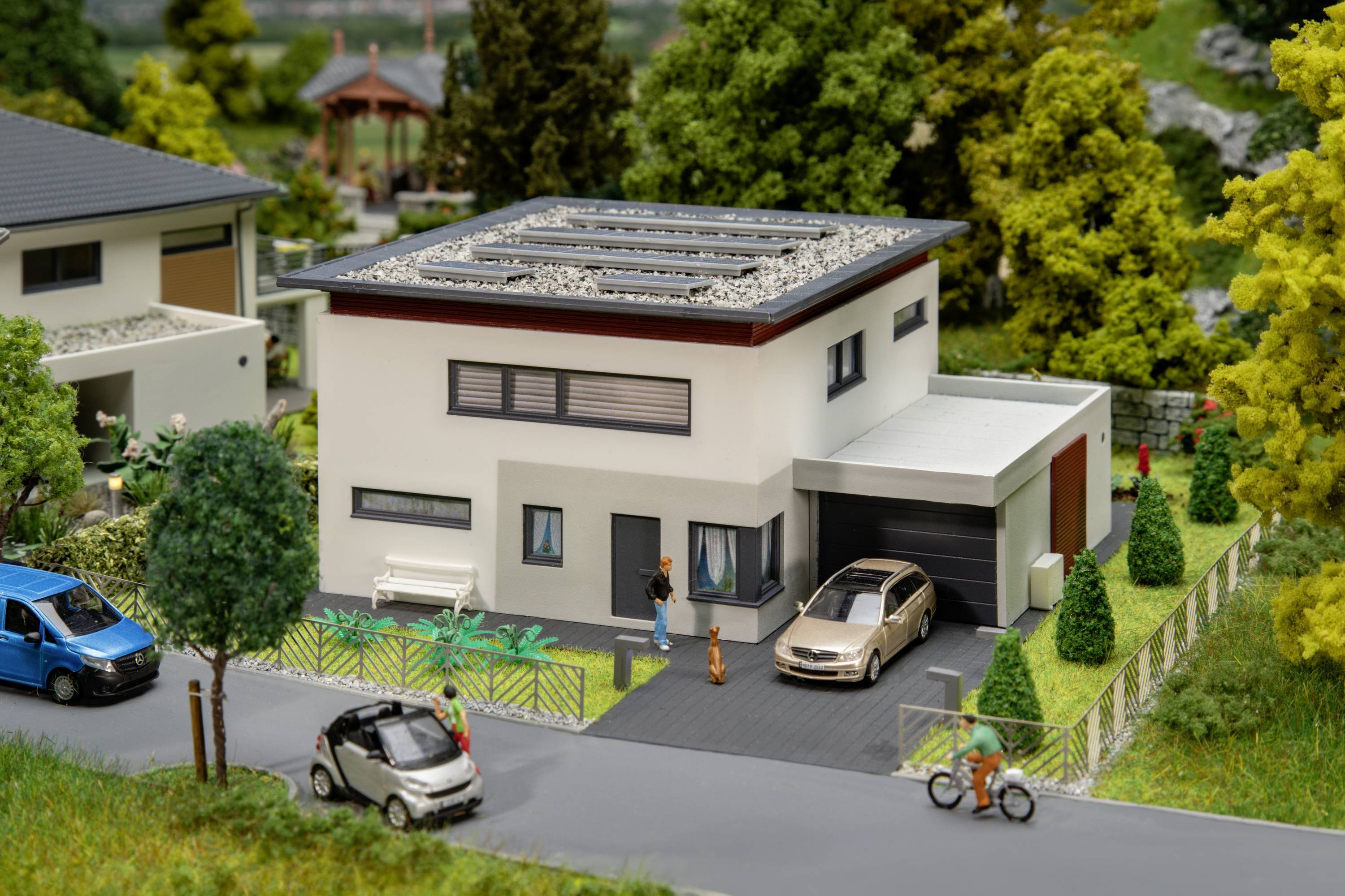Ein modernes Einfamilienhaus mit Flachdach und Solarpaneelen, umgeben von Bäumen. Eine Familie steht vor dem Haus, ein Auto parkt in der Einfahrt.