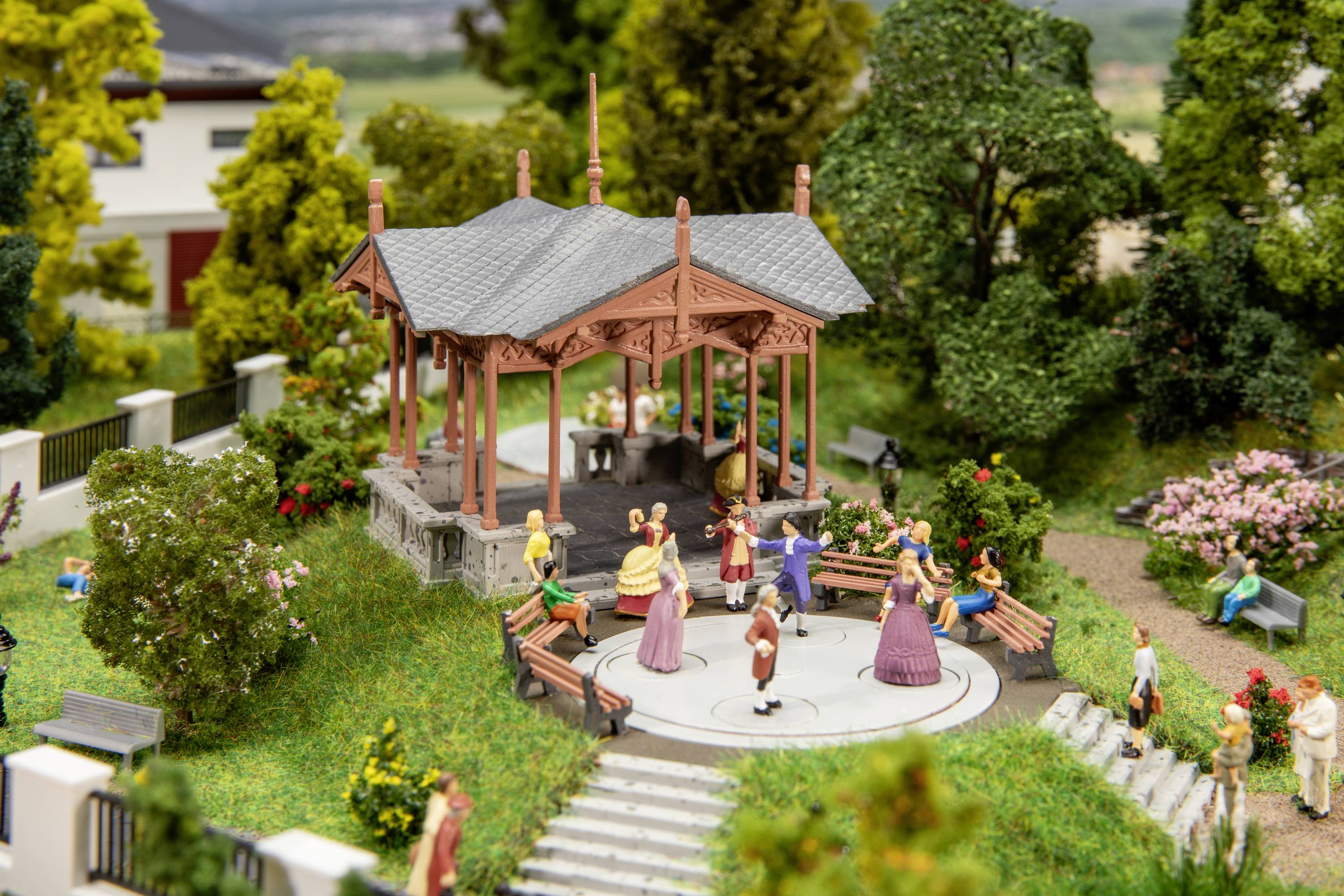 Kleine Figuren tanzen auf einer Bühne in einem Park mit Bäumen und Bänken. Es ist eine Miniaturszene mit lebendiger Atmosphäre.