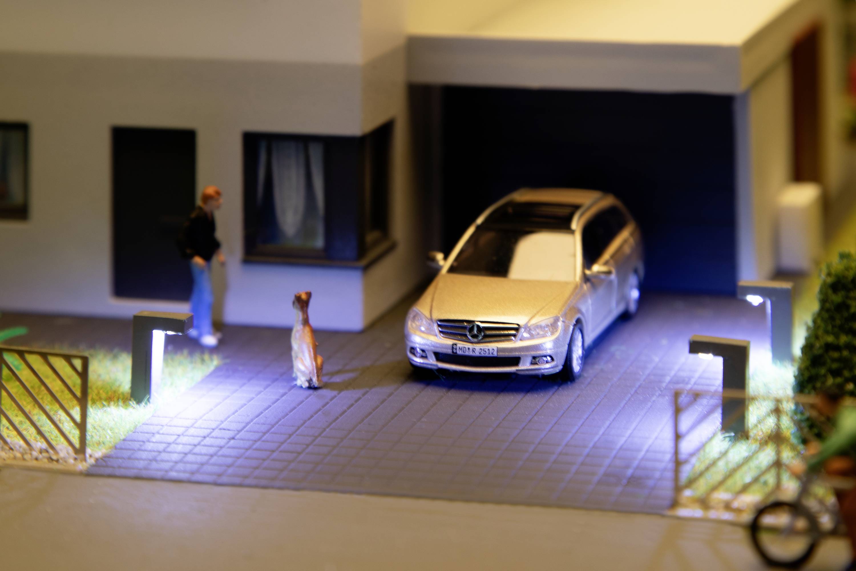 Eine Miniaturszene zeigt ein Modellauto in einer beleuchteten Einfahrt vor einer modernen Garage. Eine Figur steht neben einem Hund.