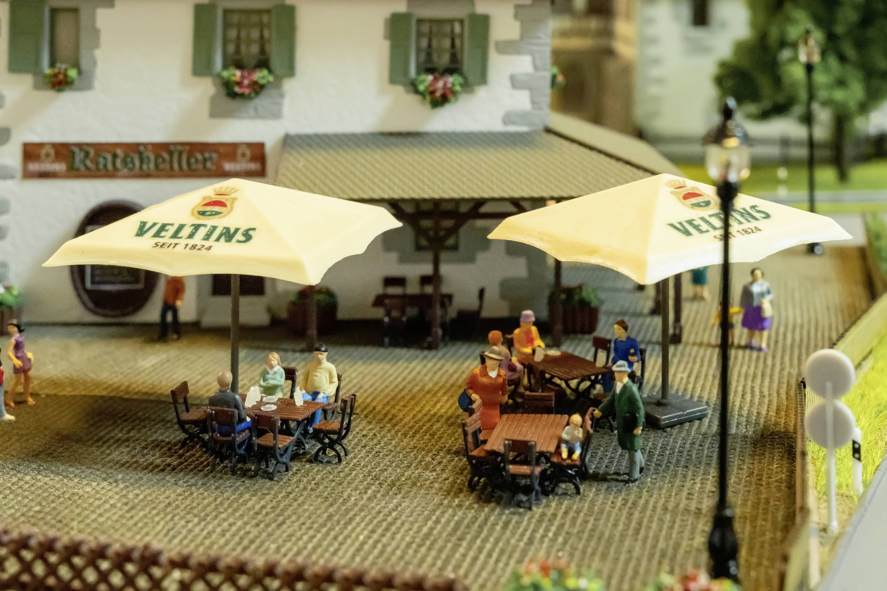 Miniatur-Straßenszene mit Menschen, die unter Außenschirmen an Tischen sitzen und ein Gebäude mit der Aufschrift 'Ratskeller' im Hintergrund.