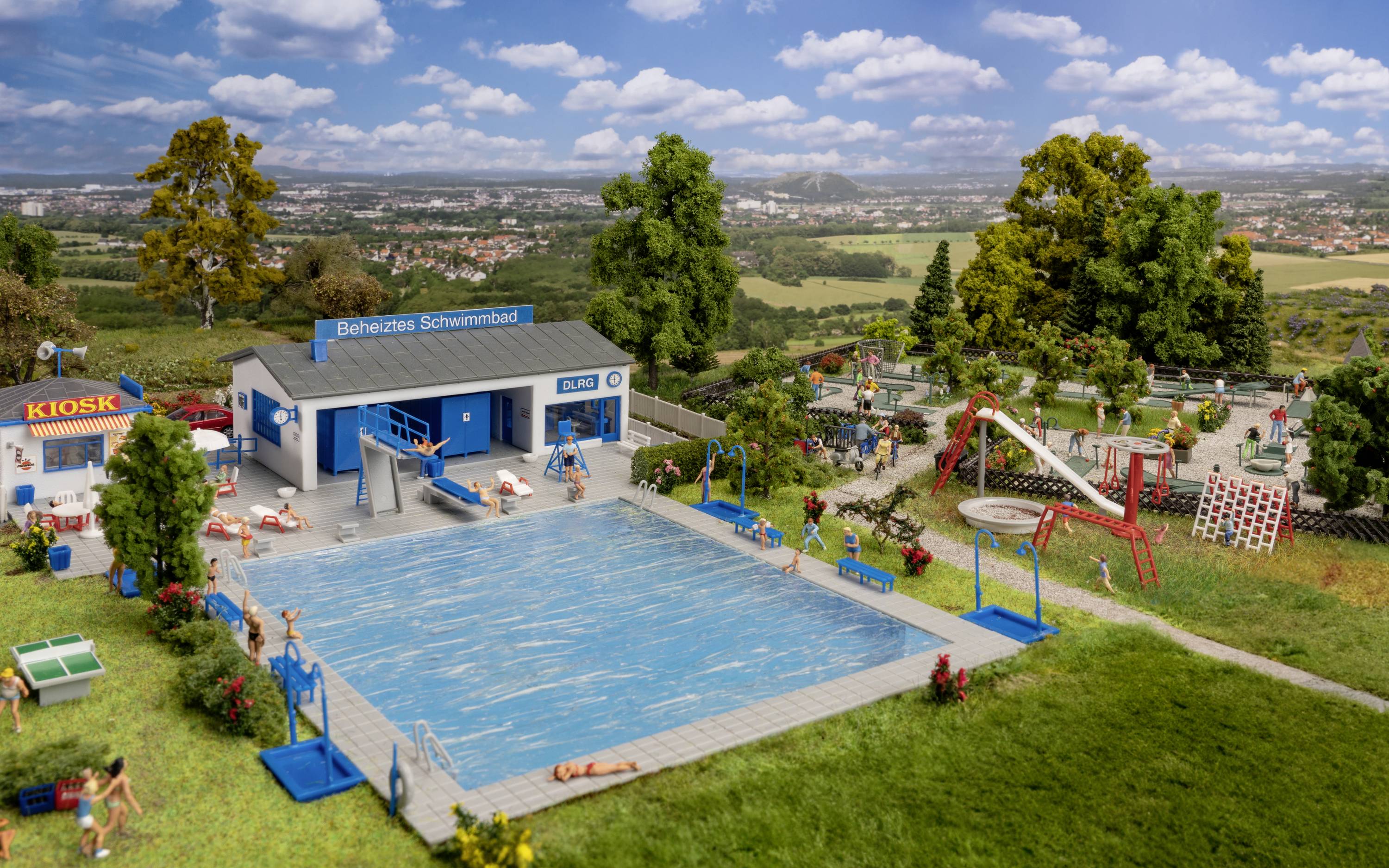Ein Modell eines Schwimmbads im Freien mit beheiztem Pool, Kiosk und Spielplatz, umgeben von Bäumen und Wiesen in einer ländlichen Umgebung.
